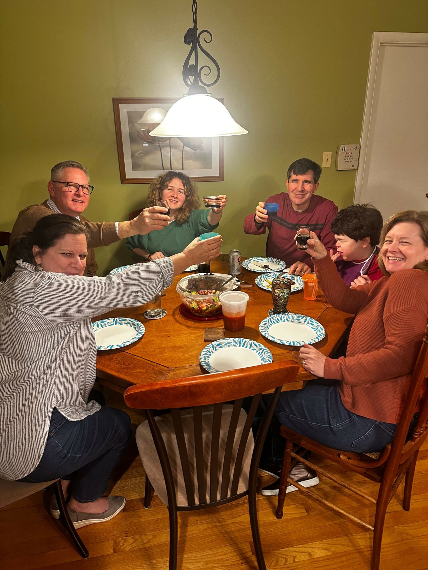 Group of five people gathered around a table with birthday cakes, plates, and drinks, celebrating indoors with a green wall and framed picture.