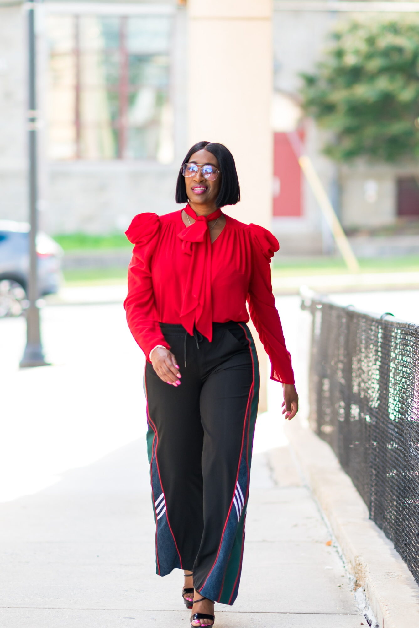 Woman walking outdoors wearing a red blouse and black pants with colorful stripes, glasses, and short hair.