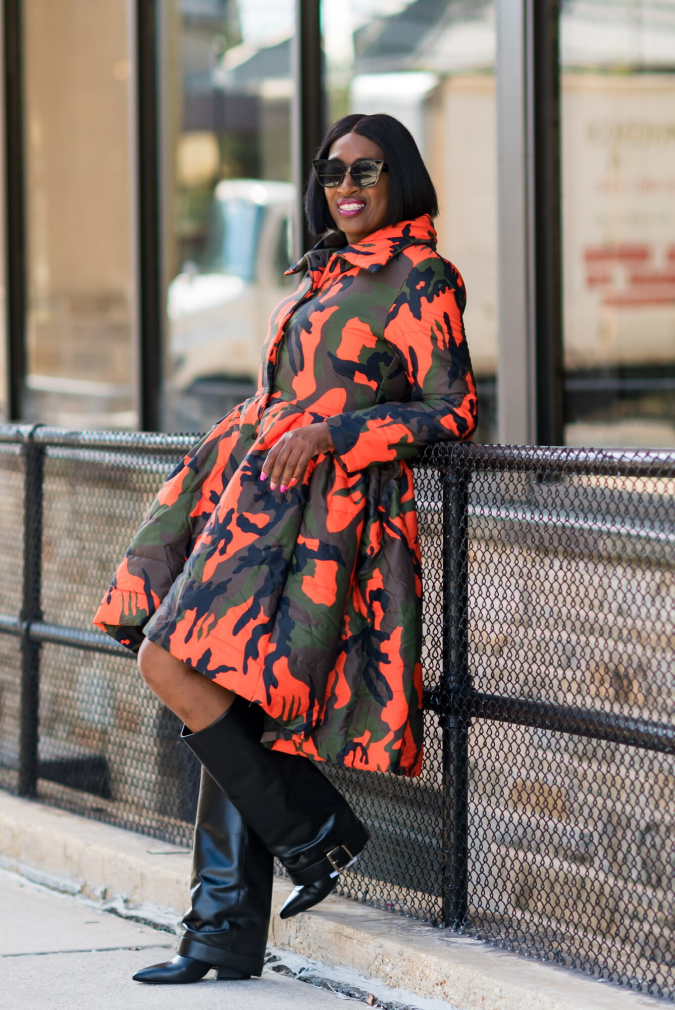 Woman in a colorful patterned coat, sunglasses, and knee-high boots leaning against a metal railing outdoors.