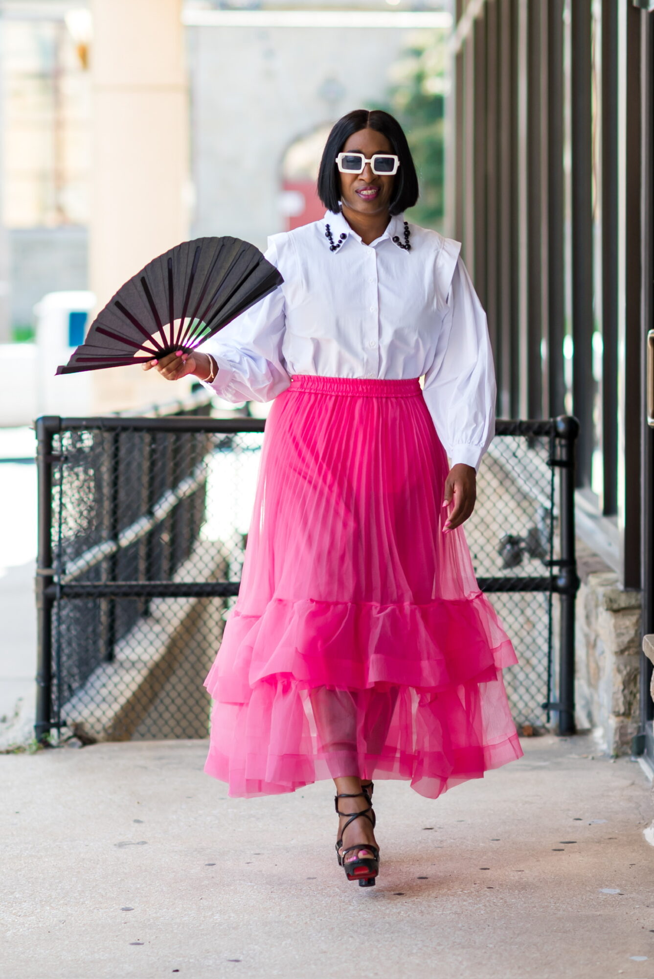 Woman walking outdoors wearing white blouse, pink tiered skirt, sunglasses, and high heels, holding a black fan.
