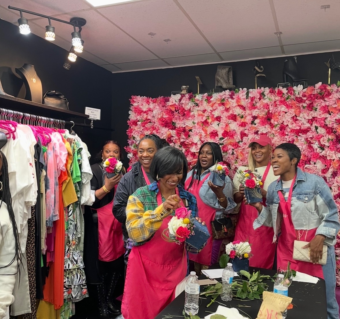 Group of women in pink aprons smiling and holding flowers in a store with a pink floral wall background.