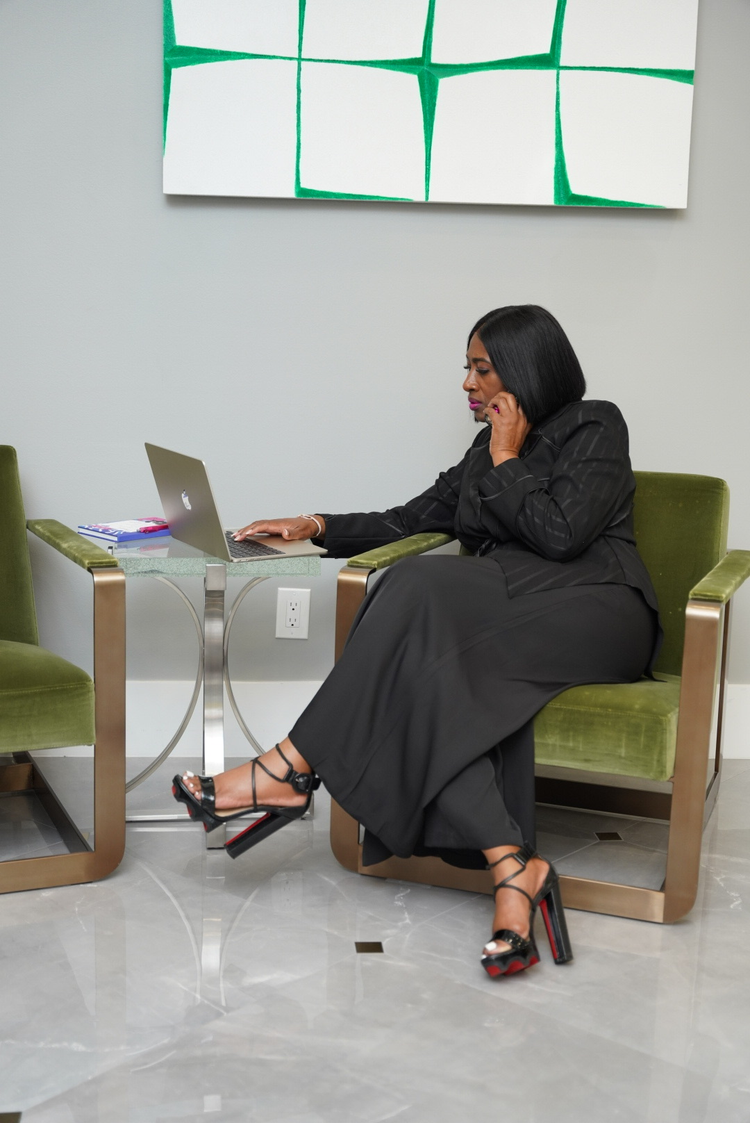 Woman sitting on a green chair using a laptop, with a white wall and abstract artwork behind her.