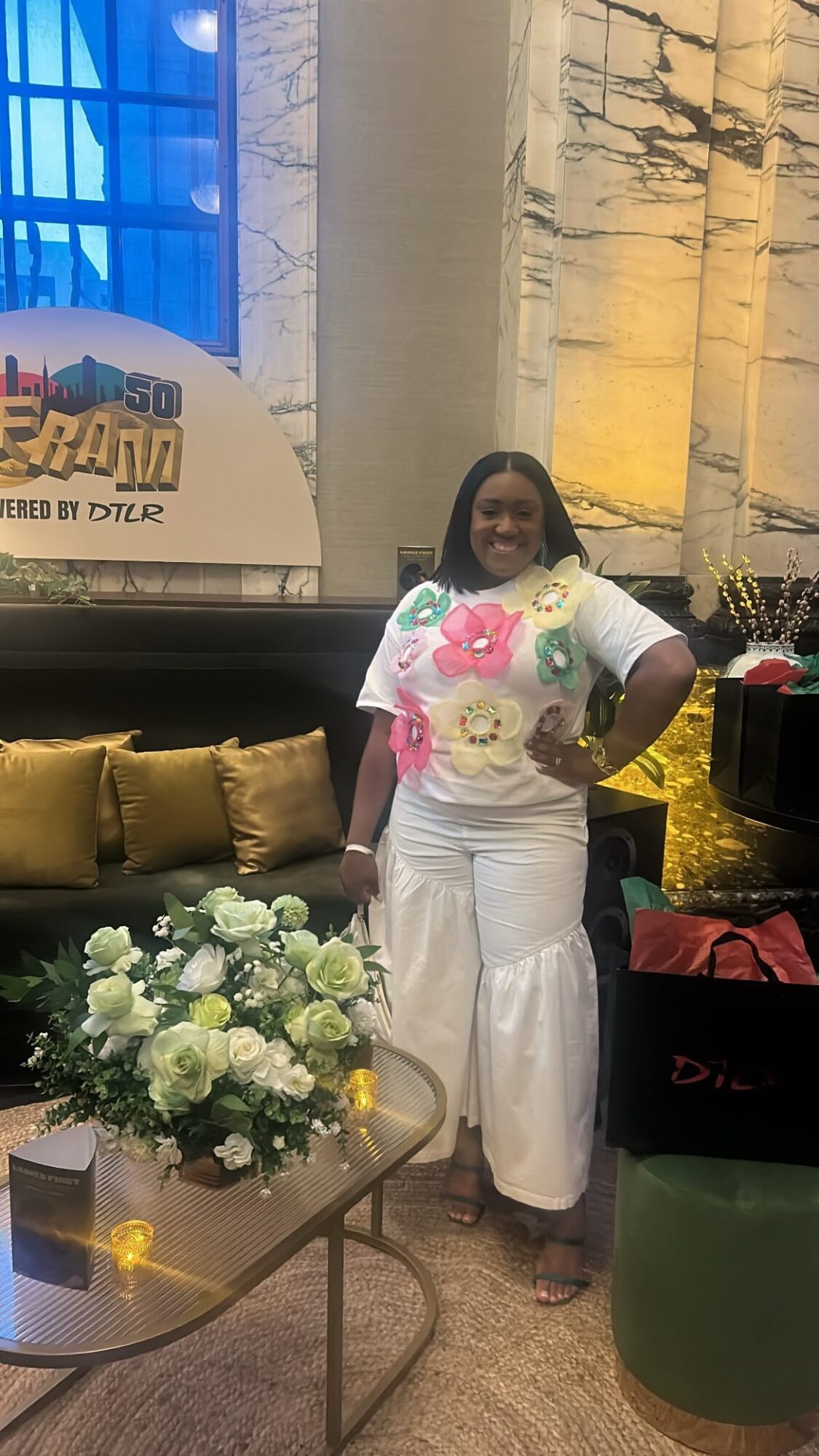 Woman standing indoors next to a table with white flowers, smiling, wearing a white dress with pink and green floral details.