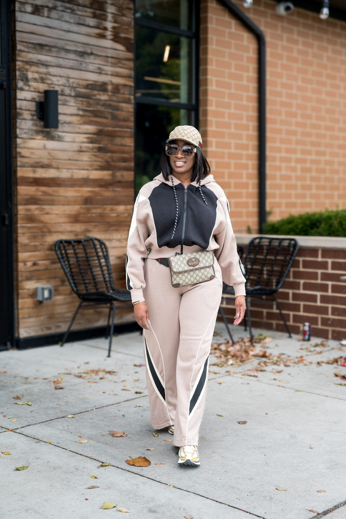 Woman in beige and black tracksuit walking outdoors, wearing sunglasses and a white cap, with chairs and brick building behind her.