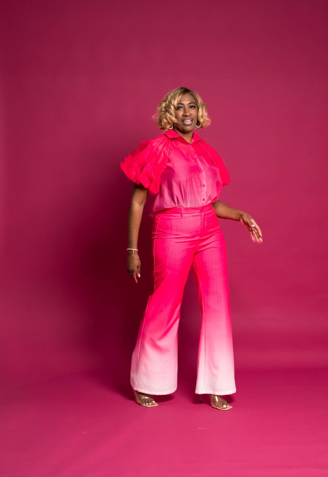 Woman with blonde hair in a pink outfit standing against a pink background, smiling, with one hand slightly raised.