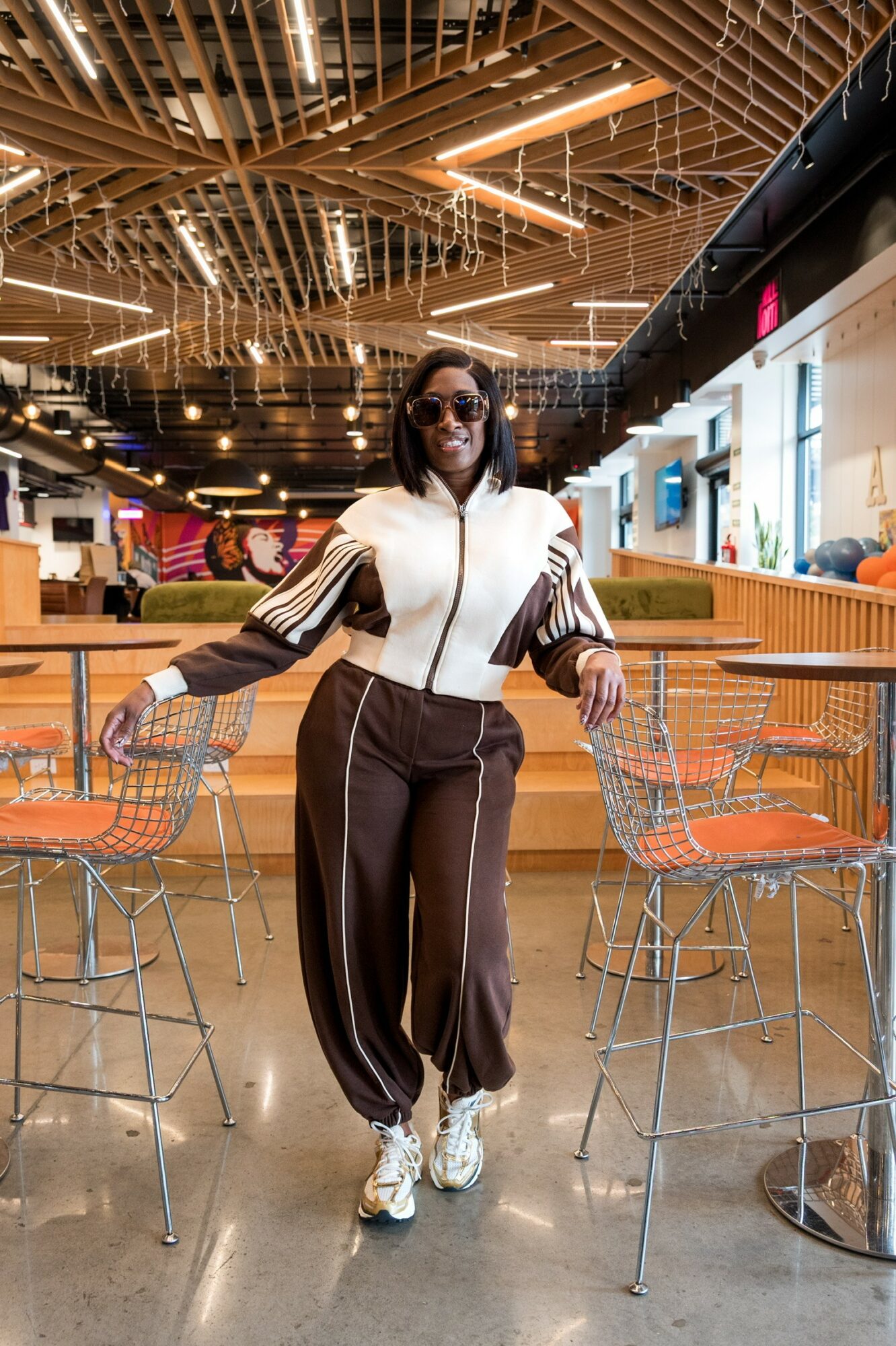 Woman in sunglasses and athletic outfit standing between two bar stools in a modern indoor space.