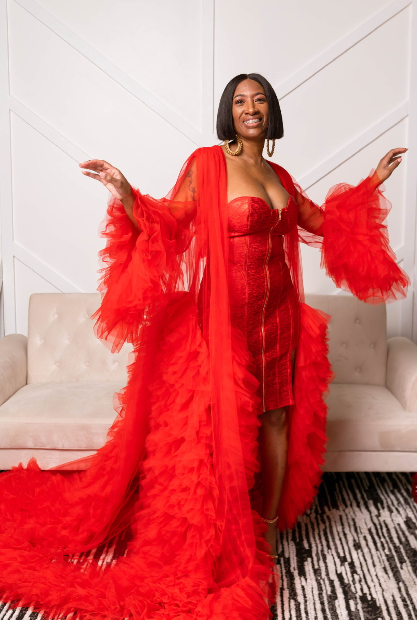 Woman in red dress and feathered cape standing in front of a sofa, smiling, arms outstretched, indoors.