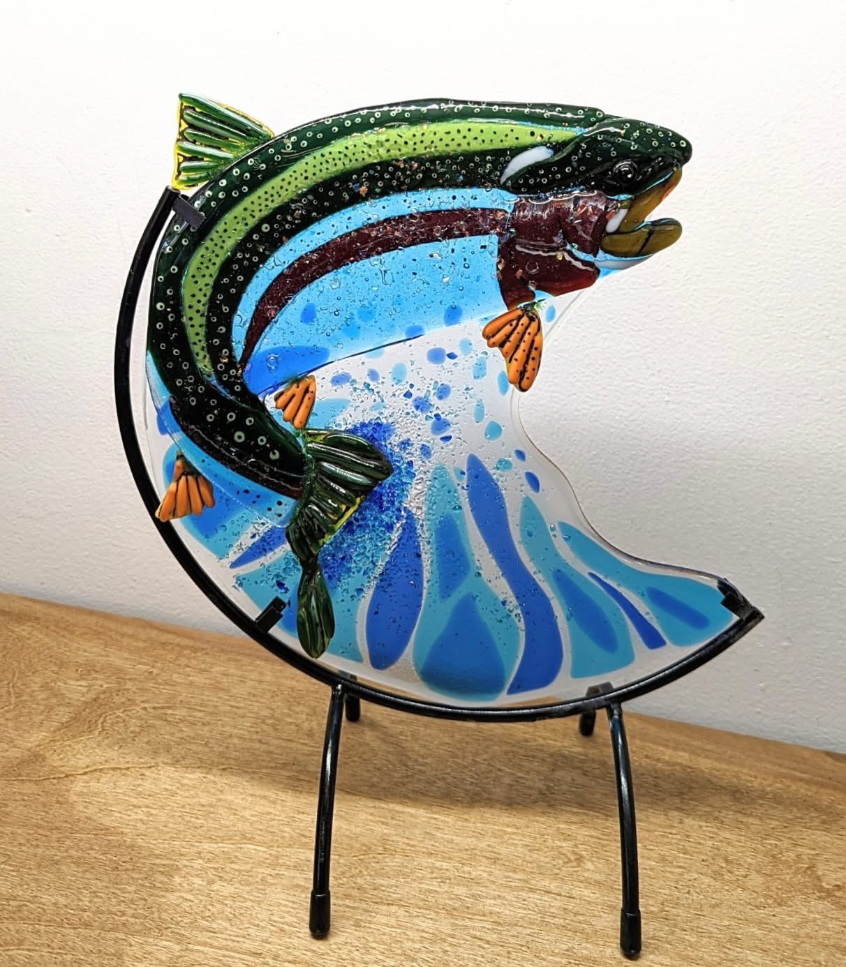 Colorful fish sculpture with a fish jumping out of water, mounted on a stand, on a wooden surface.