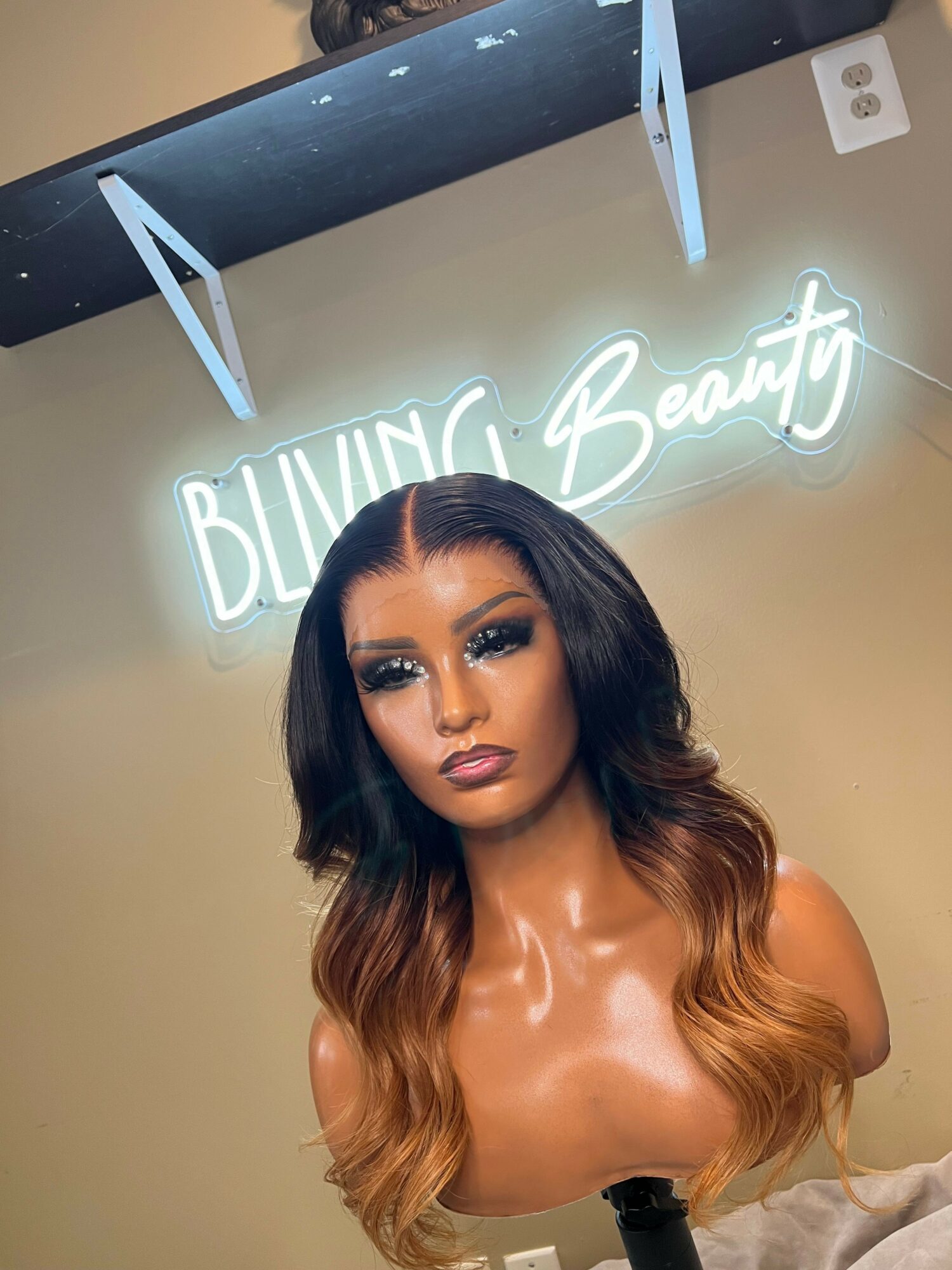 Woman with long, wavy hair and makeup, topless, in front of a beige wall with a neon sign reading 'Beauty'.