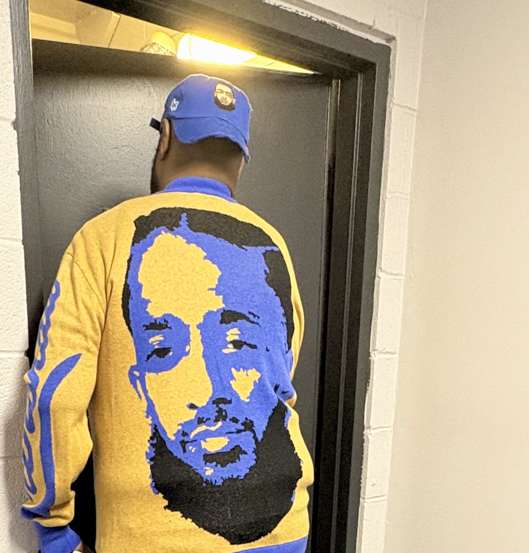 Person wearing a blue cap and a yellow and blue sweater with a face design, standing in front of a door.