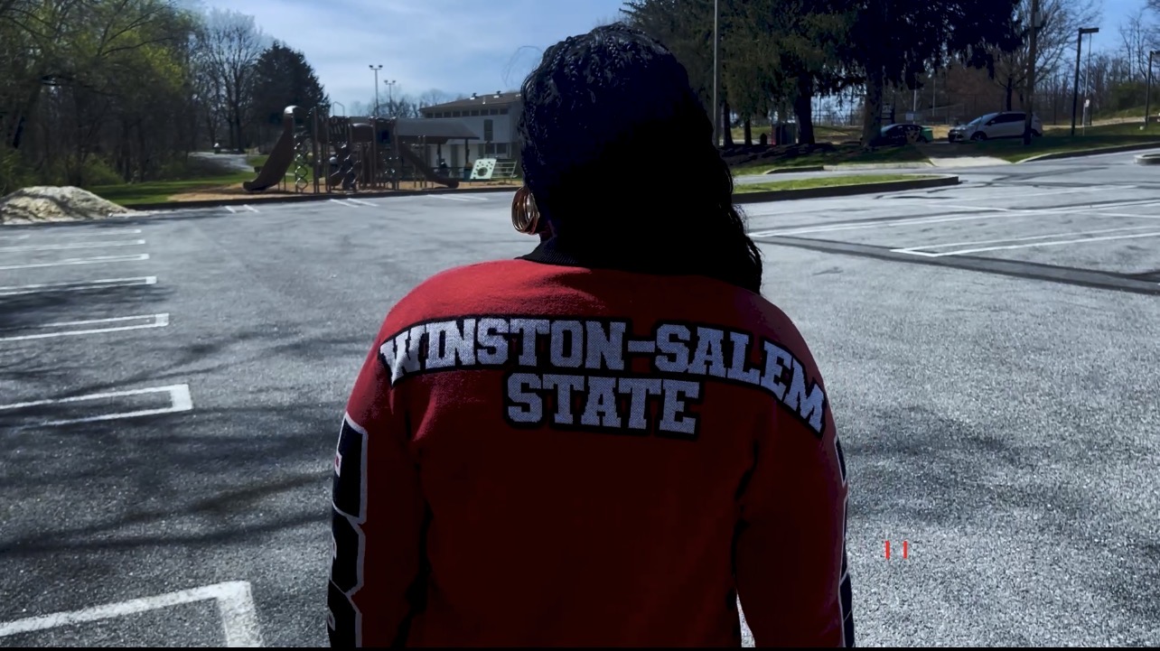 Person with dark hair wearing a red jacket with white and black text on the back, standing in an empty parking lot.