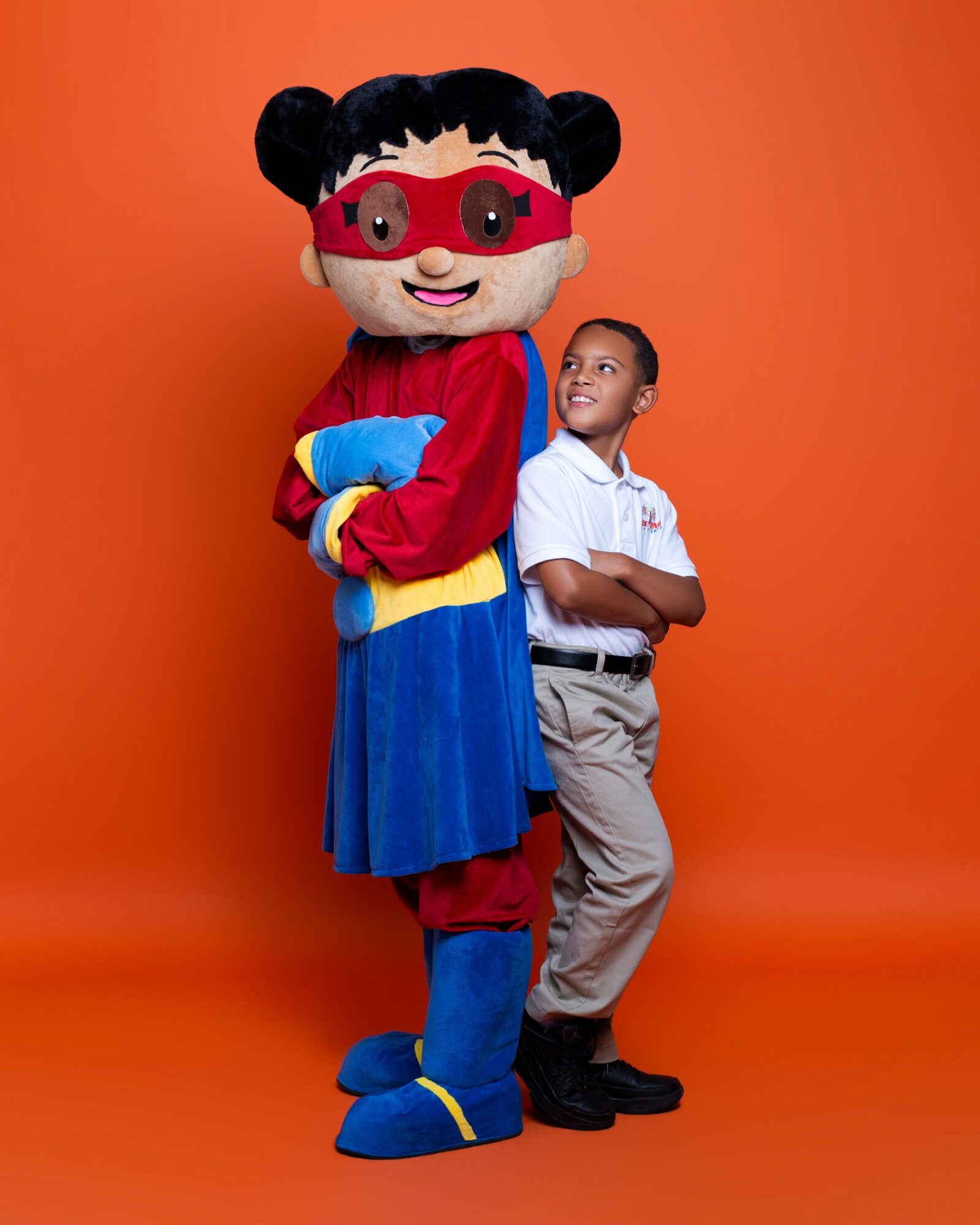 Child standing with arms crossed next to a large mascot character with black hair, red mask, and colorful costume, against orange background.