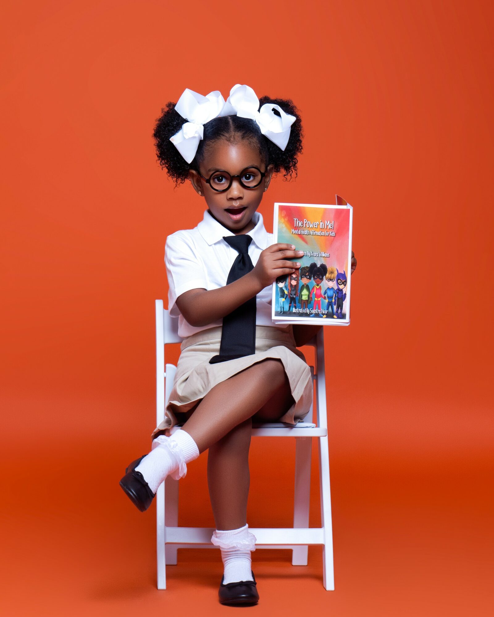 Young girl with glasses and large white bows in her hair, sitting on a white chair, holding a colorful book, against an orange background.