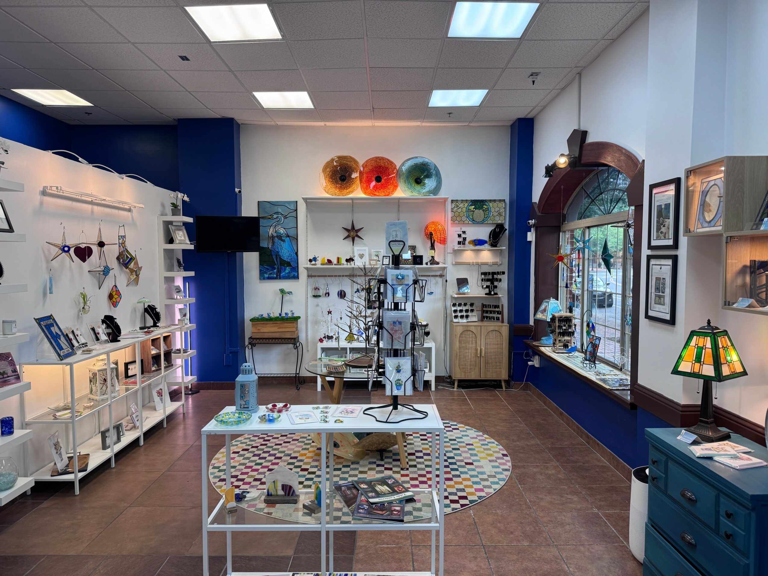 Interior of a store with shelves, colorful glass art, and a table with a rainbow rug, decorated with various items.