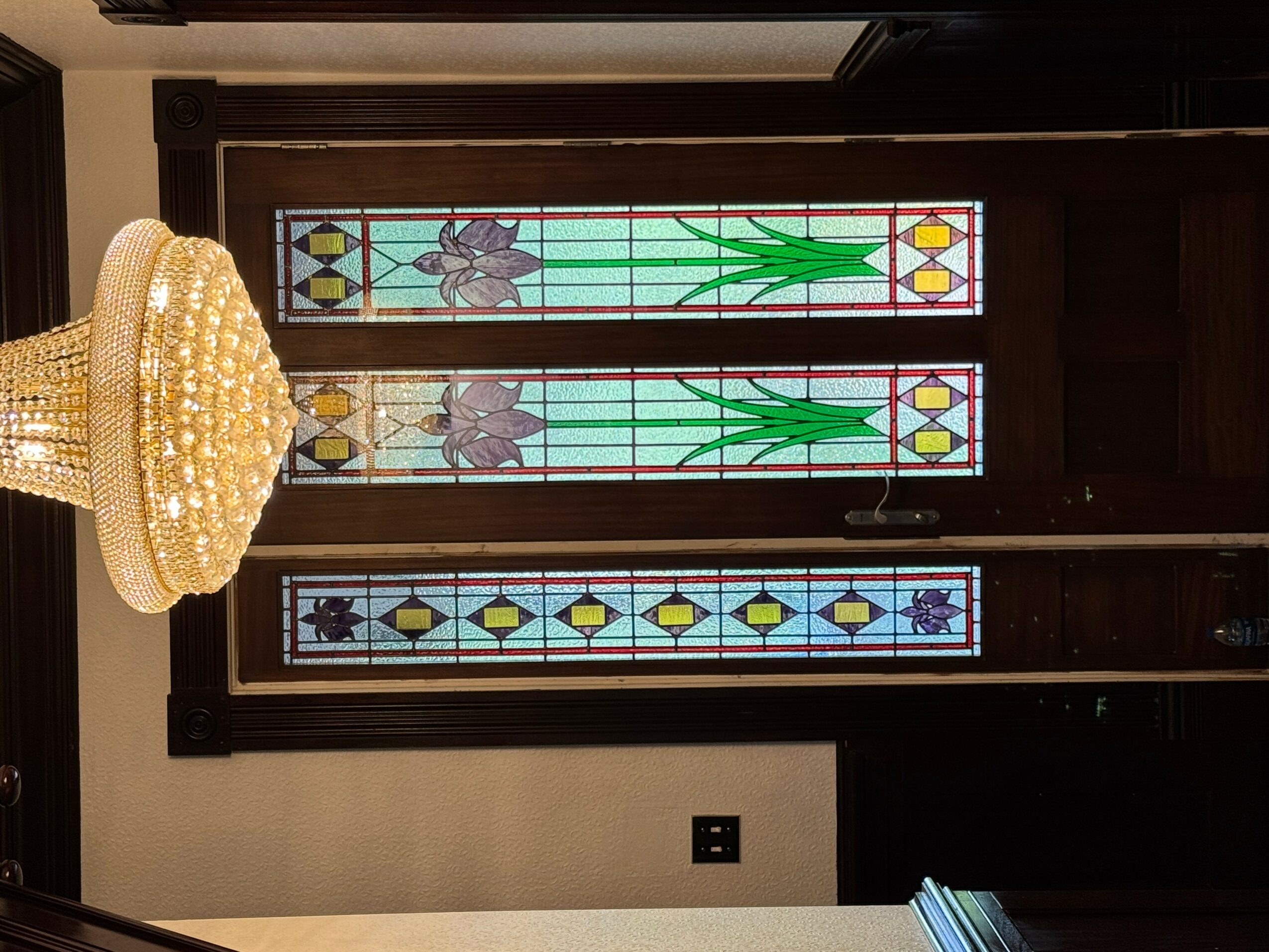 Colorful stained glass window with floral and geometric patterns, seen through a dark wooden frame, with a chandelier in front.