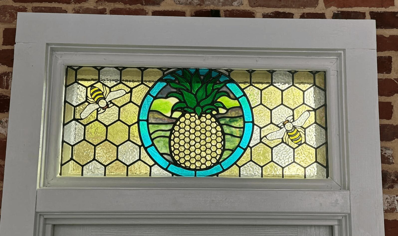 Decorative stained glass window with a pineapple, honeybees, and honeycomb pattern, framed by white trim, against a brick wall.
