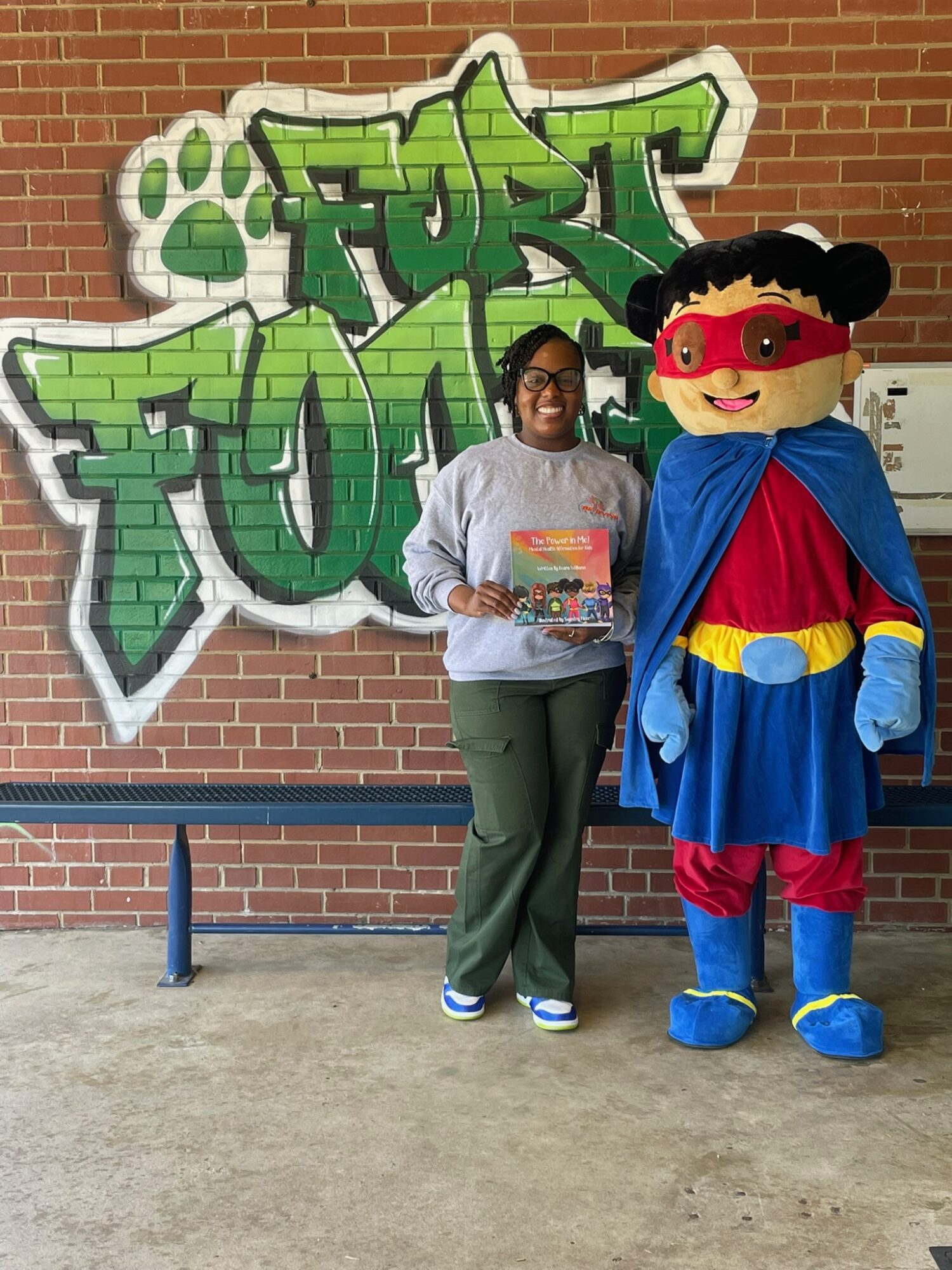Person standing next to a mascot in front of graffiti on a brick wall, holding a colorful book.
