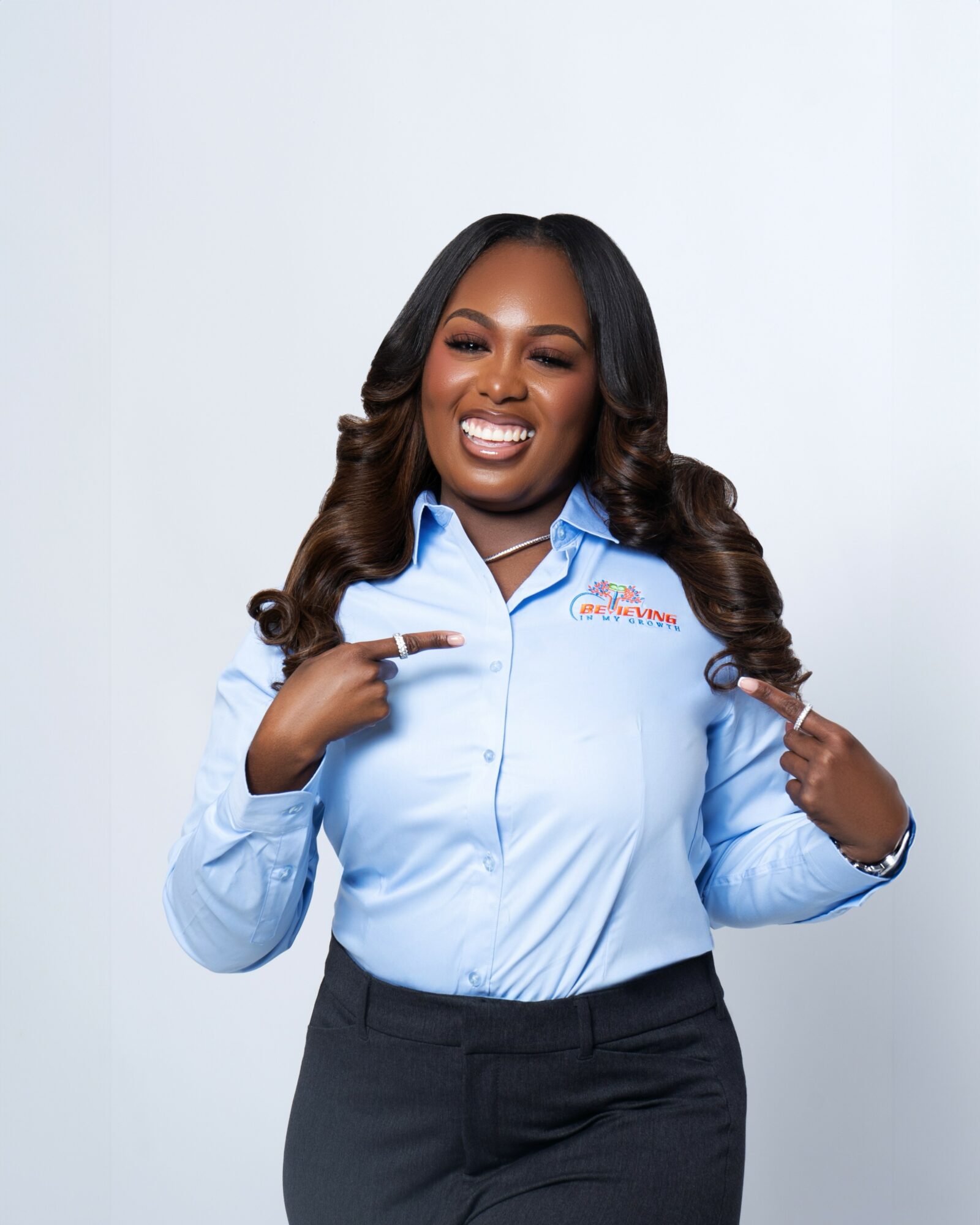 Smiling woman with long wavy hair pointing to her shirt, standing against a plain background.