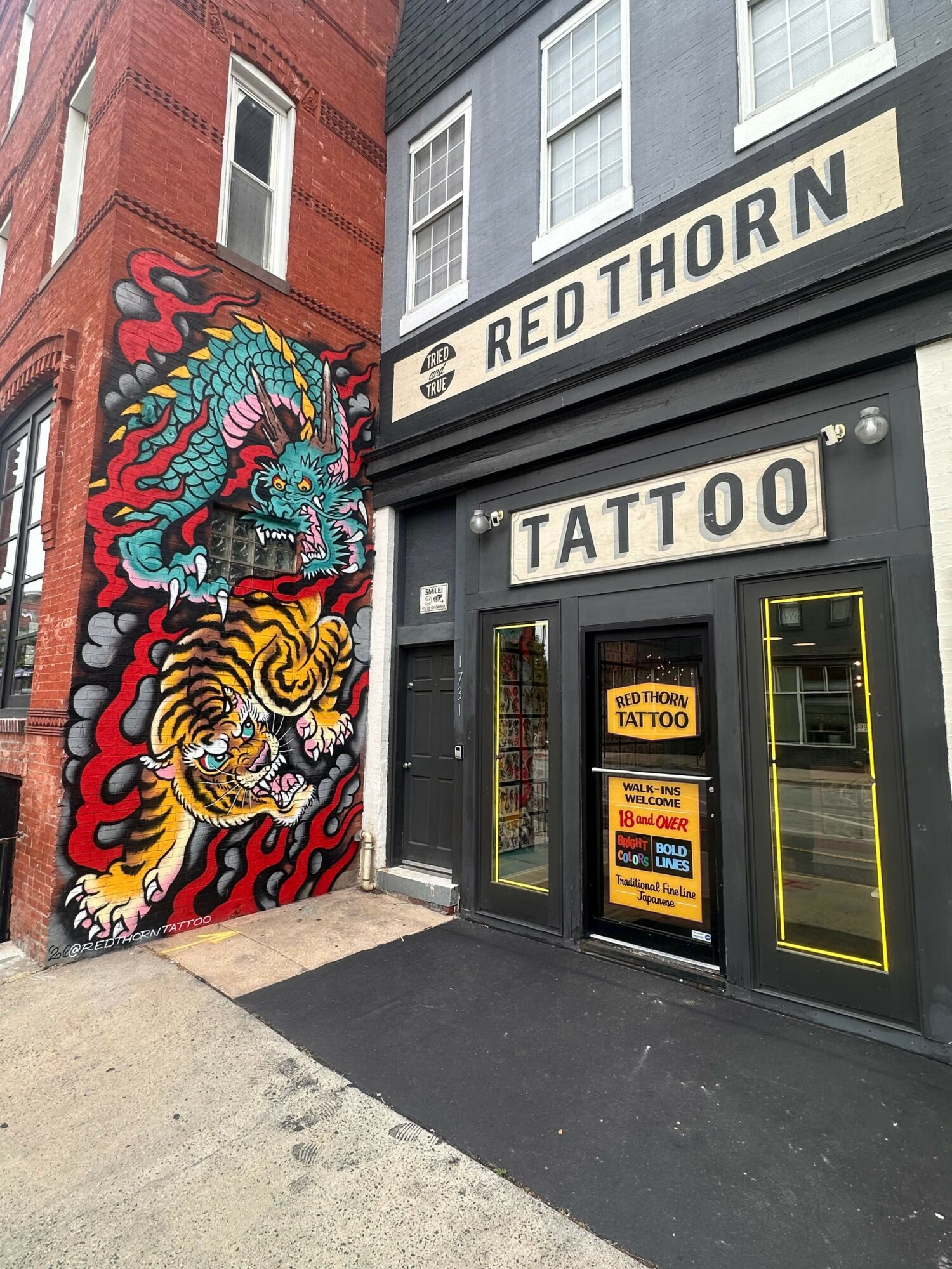 Tattoo shop entrance with colorful dragon and tiger mural on wall, sign for tattoo and piercing, and storefront windows.