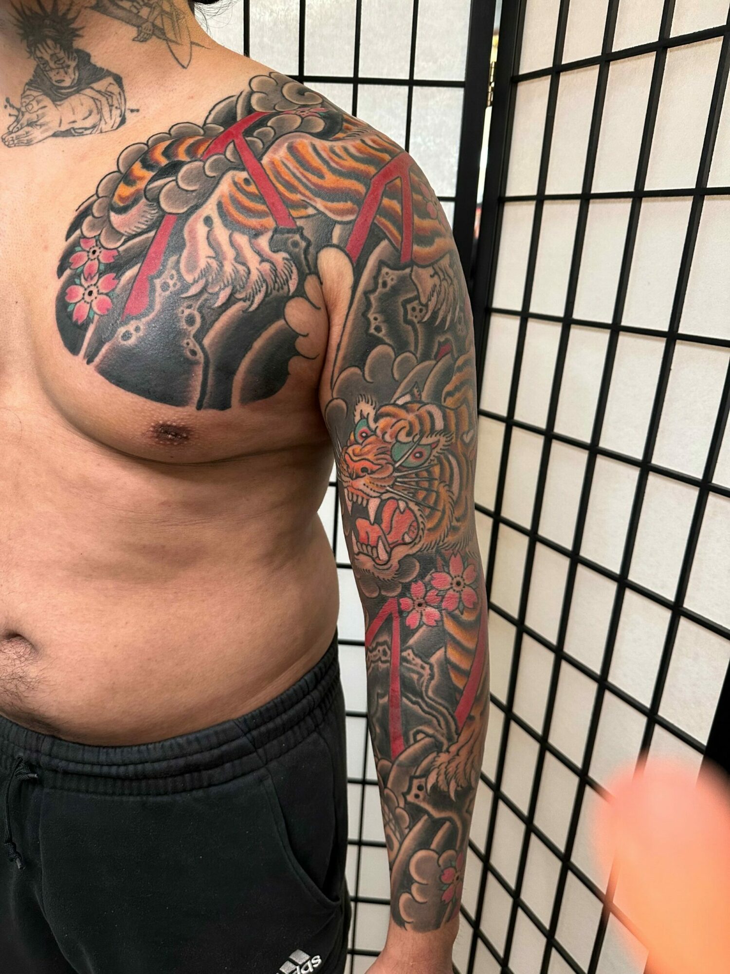 Tattooed arm resting on a black grid surface, with part of the torso visible, wearing black pants.
