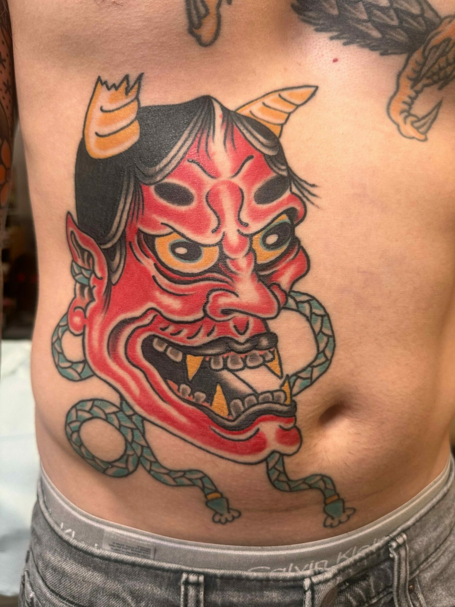 Tattoo of a red demon face with horns, sharp teeth, and a snake coiled around it on a person's side.