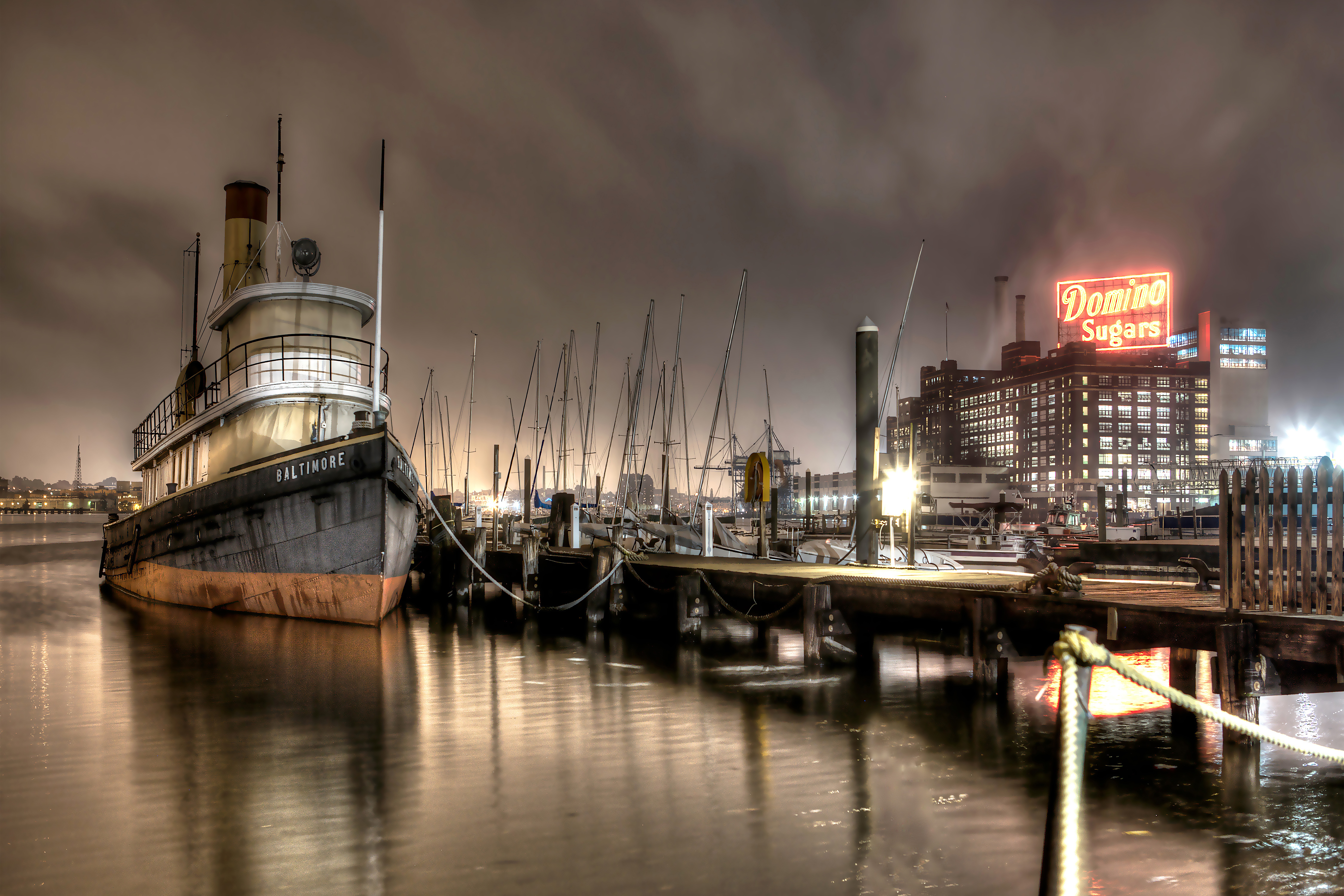 A docked boat at night with a city skyline and illuminated sign in the background, reflecting on calm water.