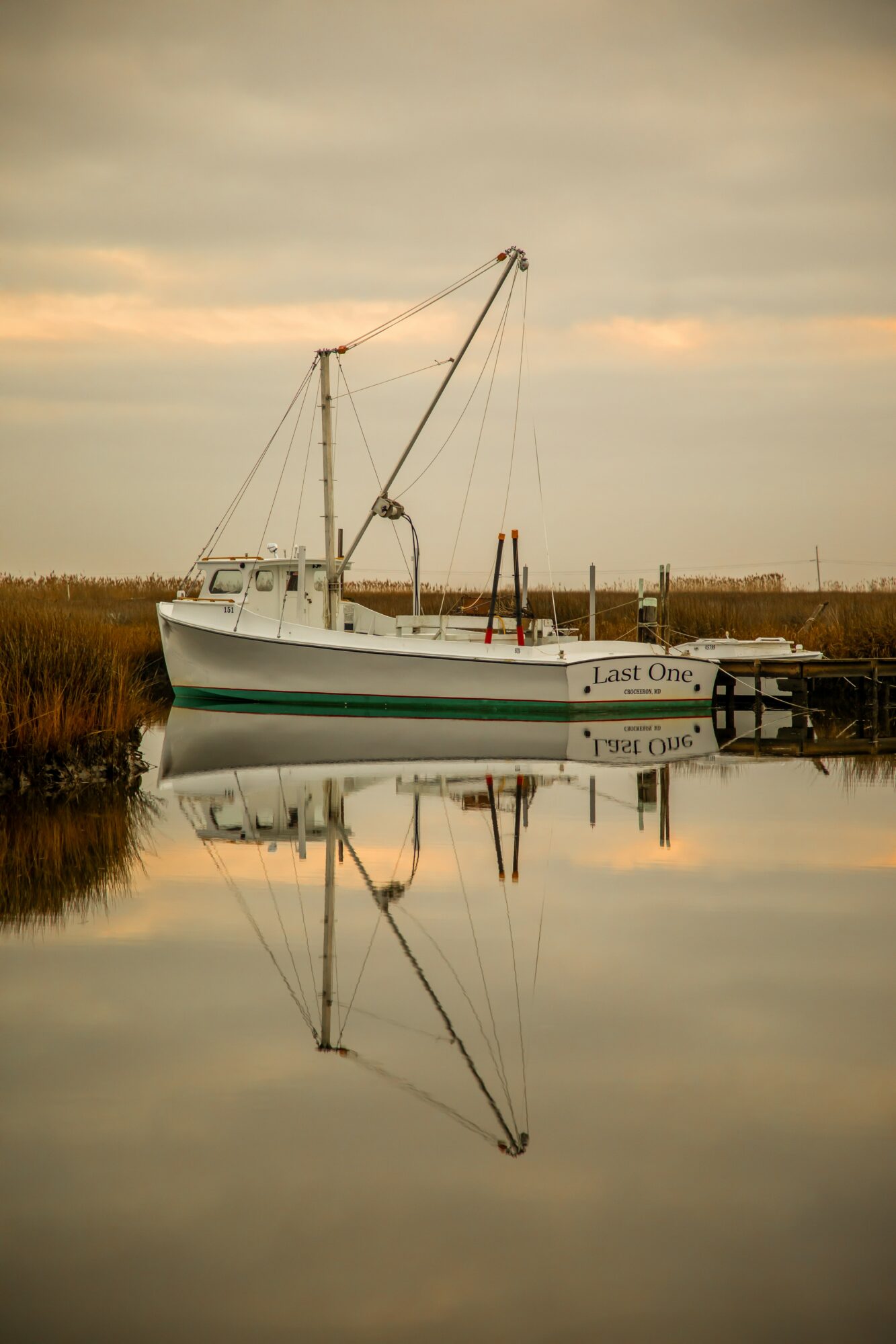 Sailboat docked near marshland with calm water reflecting the boat and sky, during sunset or sunrise with cloudy sky.