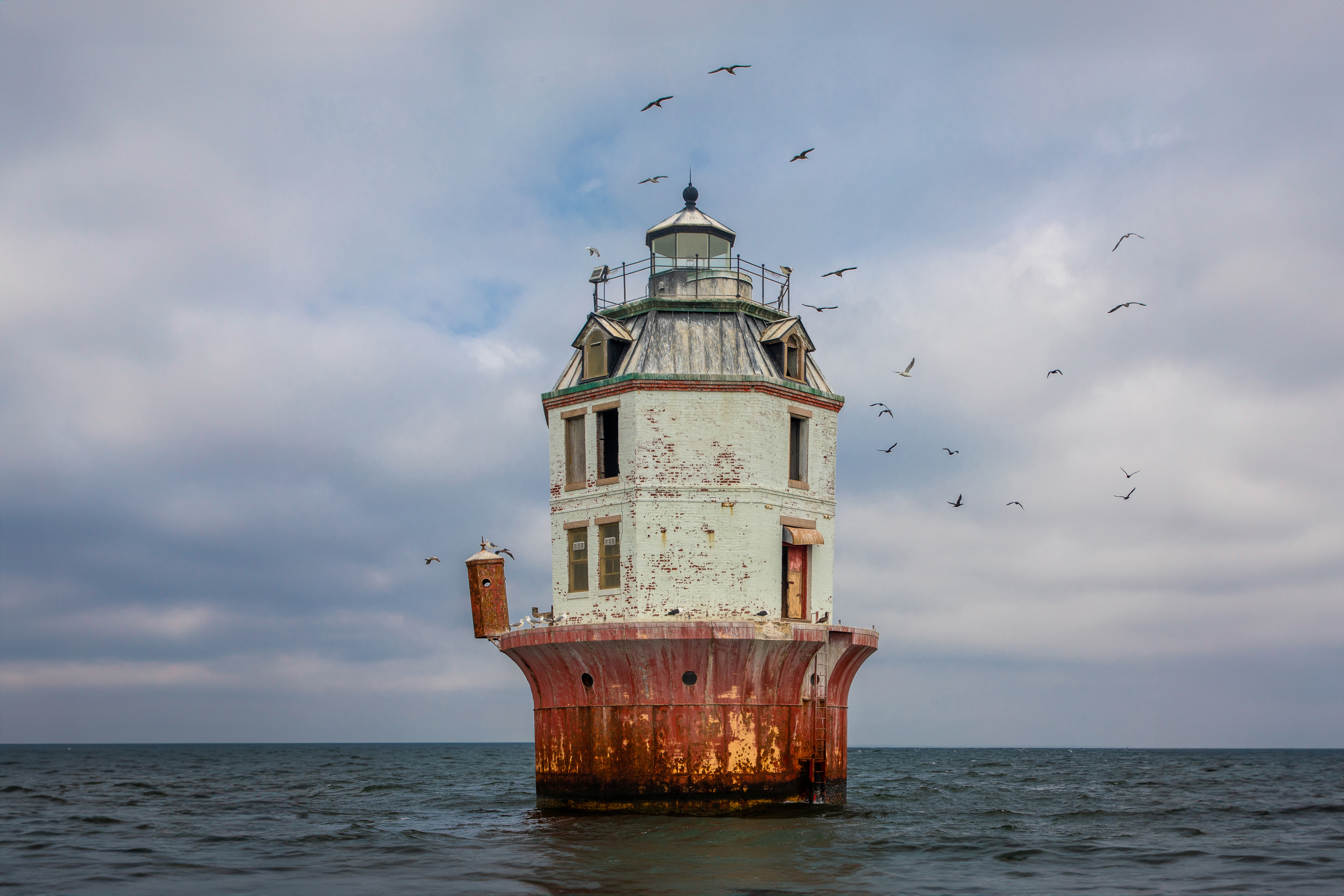A lighthouse standing in the water with a cloudy sky and birds flying around it.