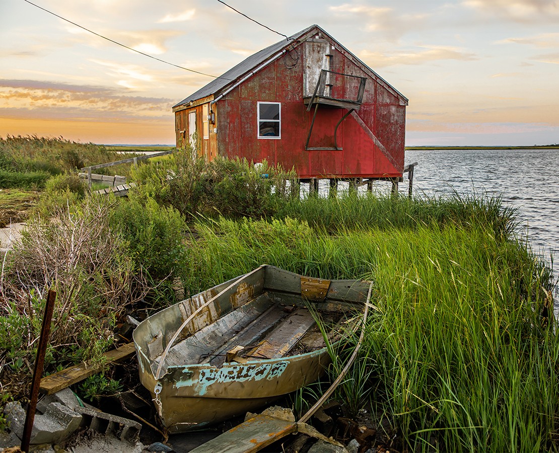 Old red wooden house on stilts near water, with an abandoned boat in tall grass in foreground, cloudy sky.