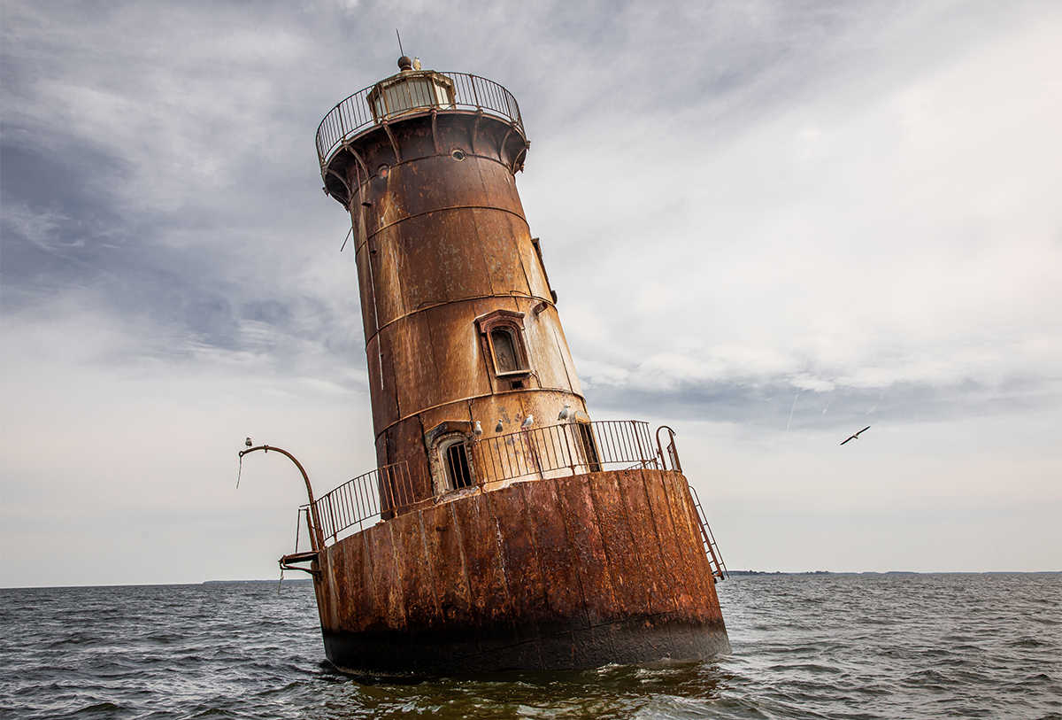 Rusty, tilted lighthouse leaning in water with cloudy sky background, railing around top, and a bird flying nearby.