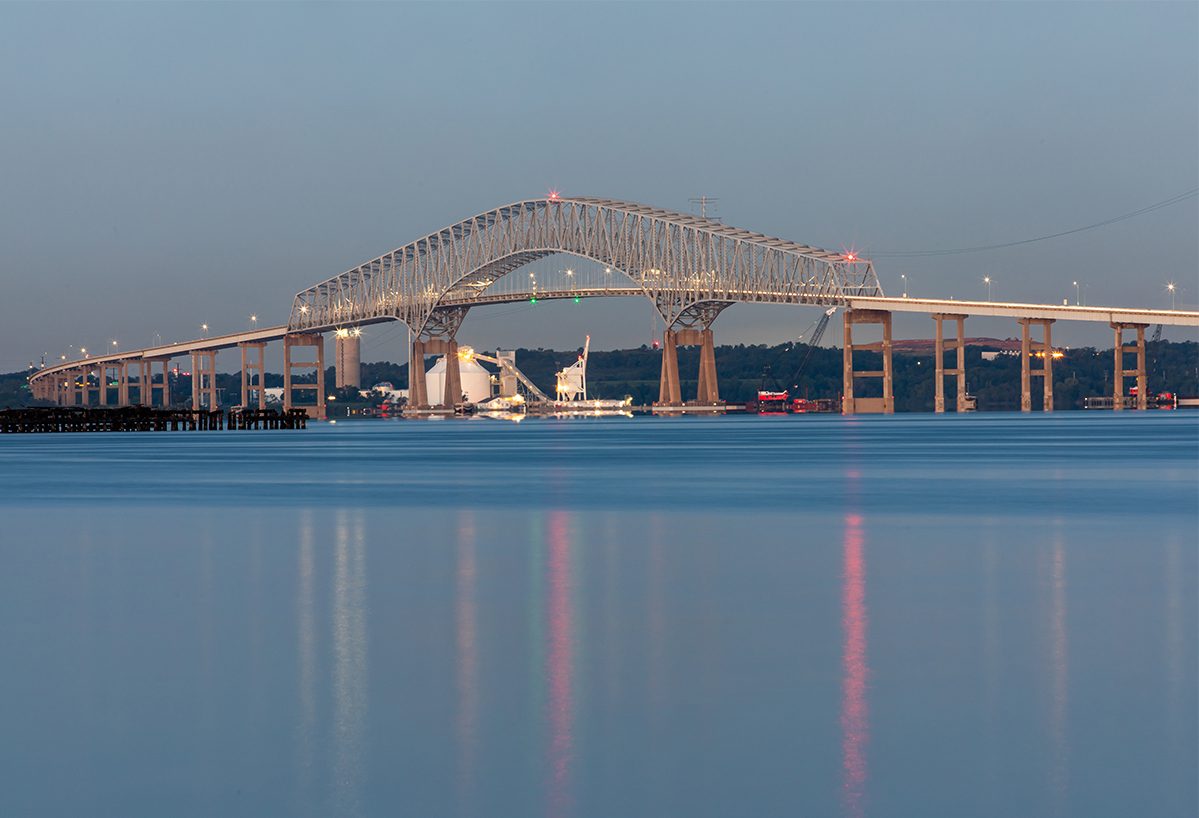 Bridge over water with city skyline in the background, under a clear sky, reflected in the water.