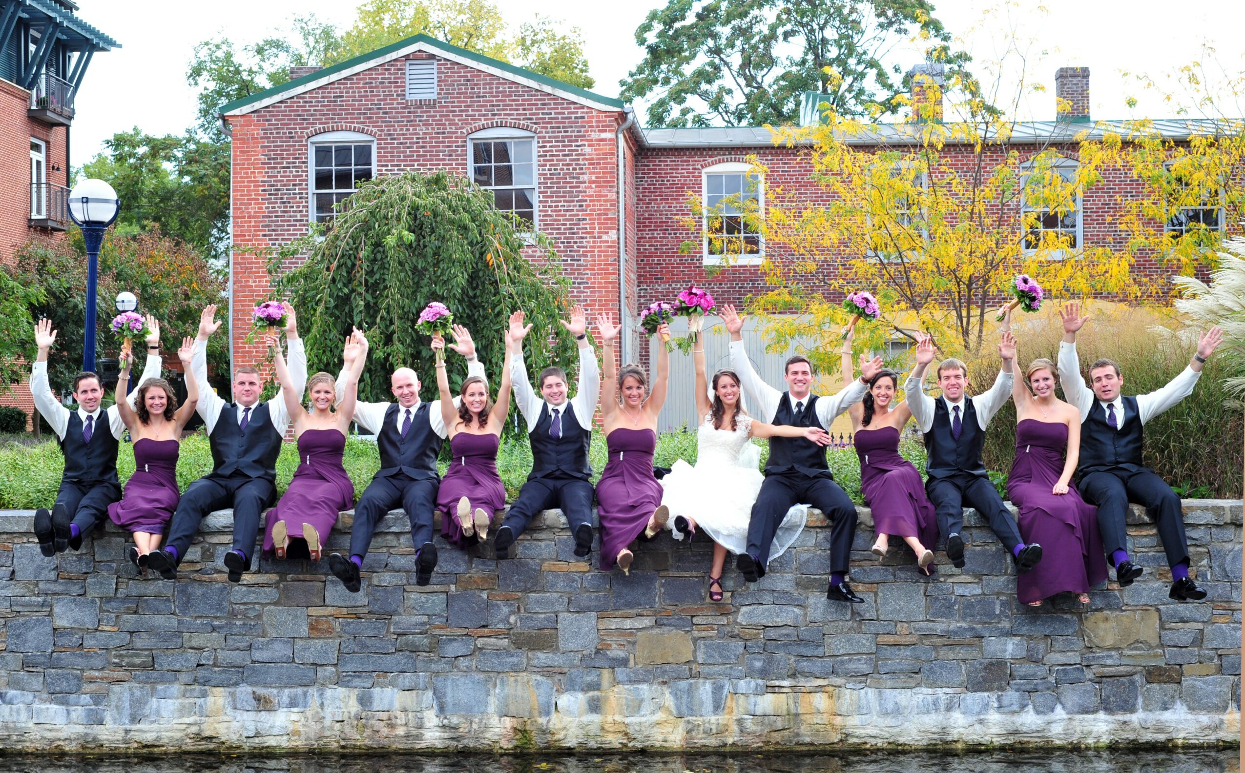 Group of people in formal attire sitting on a stone wall, raising their hands outdoors with buildings and trees in background.