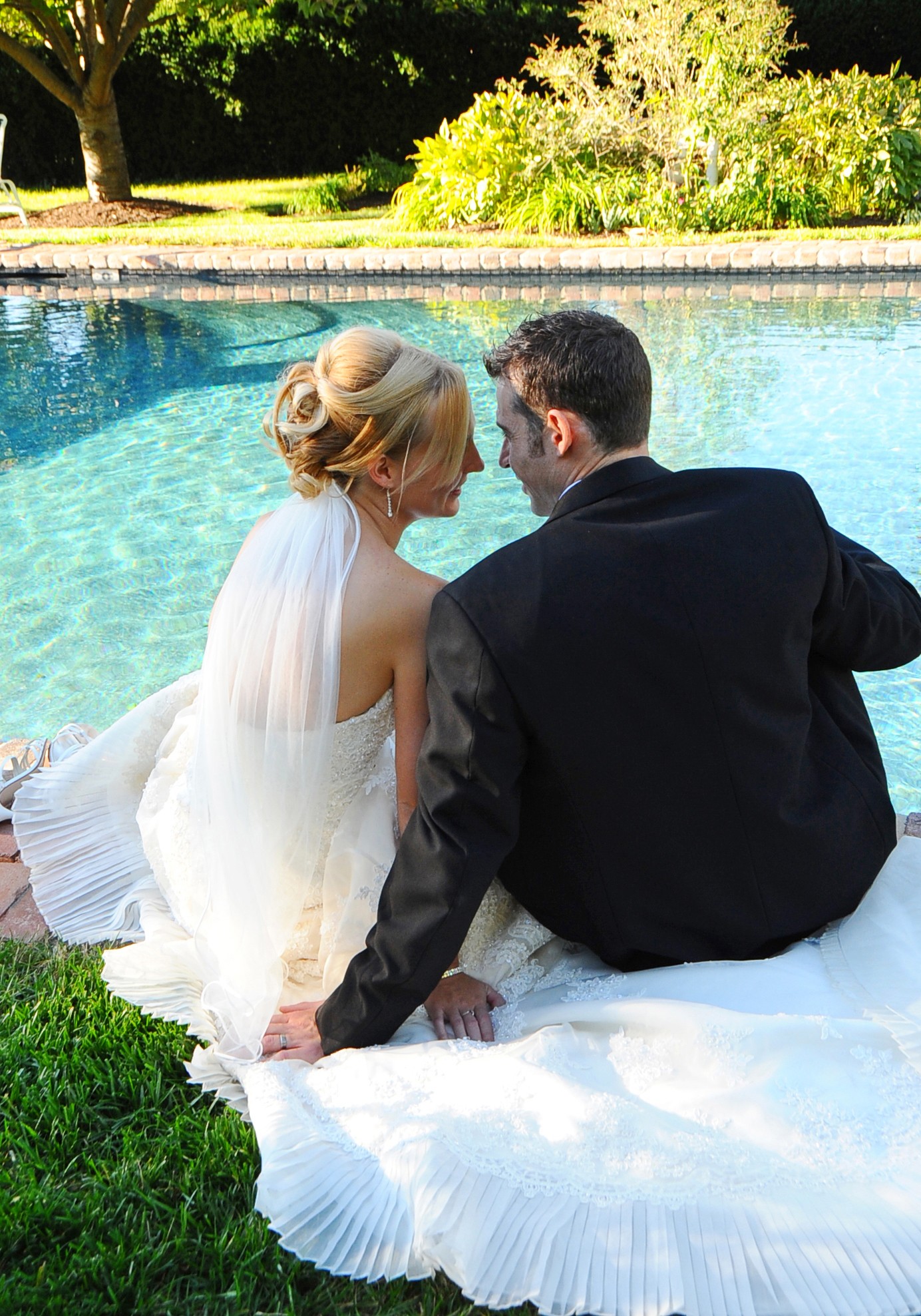 Bride and groom sit by a pool, holding hands, with trees and bushes in the background.