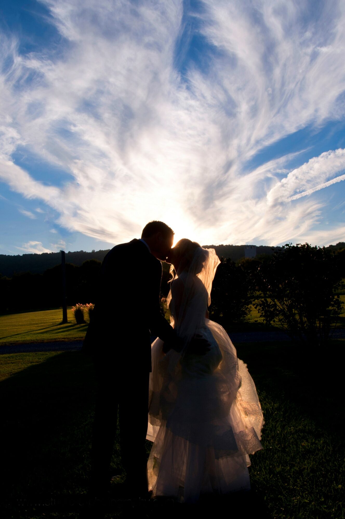 Silhouette of a couple kissing outdoors with a bright sky and clouds behind them, sunlight creating a glow around them.