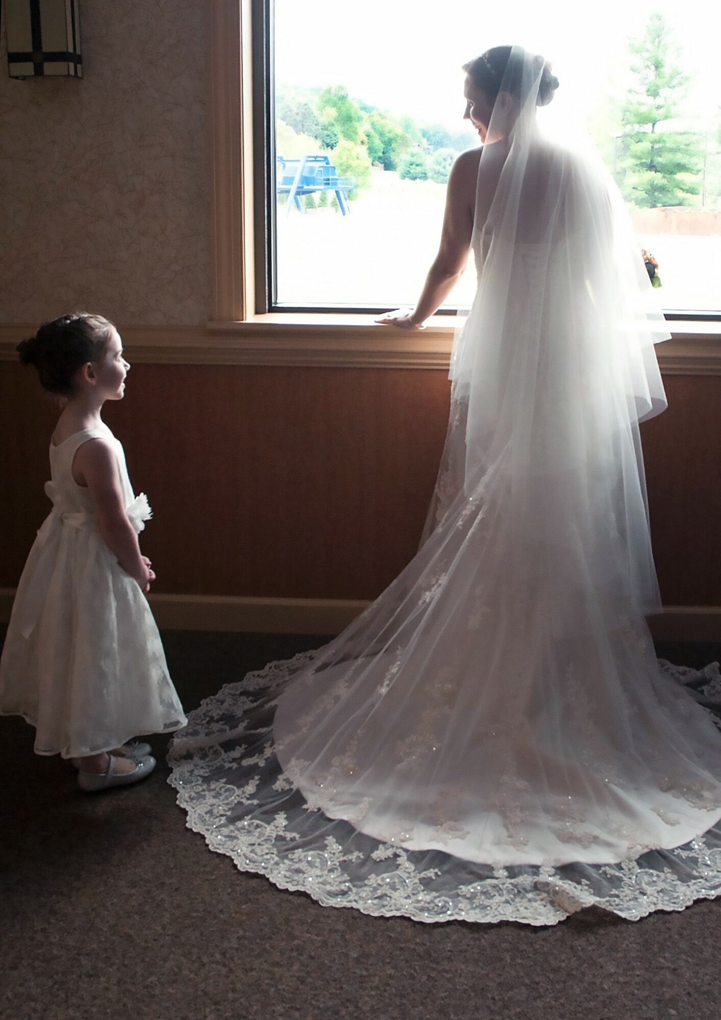 A woman in a wedding dress looks out a window, a young girl in a dress stands nearby inside a room.