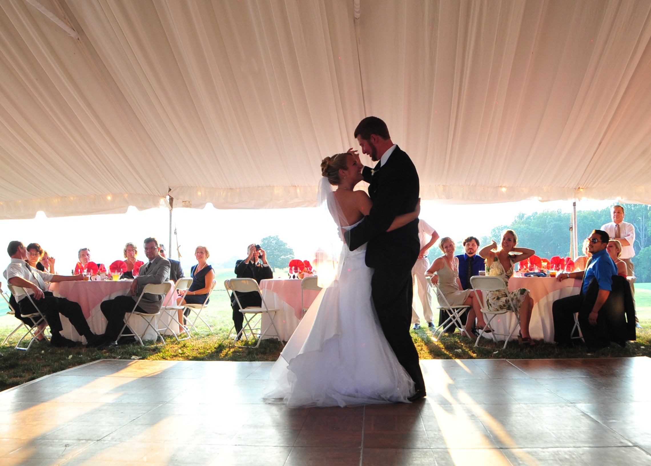 Couple dancing under a large tent with seated guests at tables in the background.