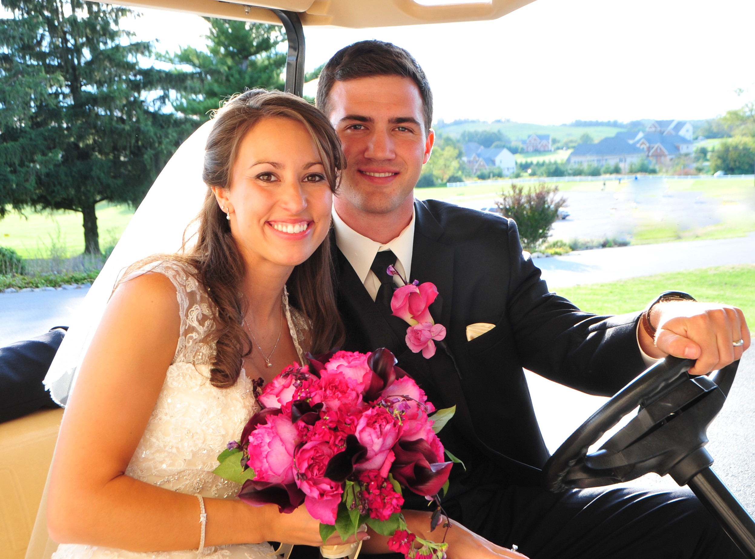 Bride and groom sitting in a vehicle, smiling, with a scenic outdoor background, bride holding a pink bouquet.
