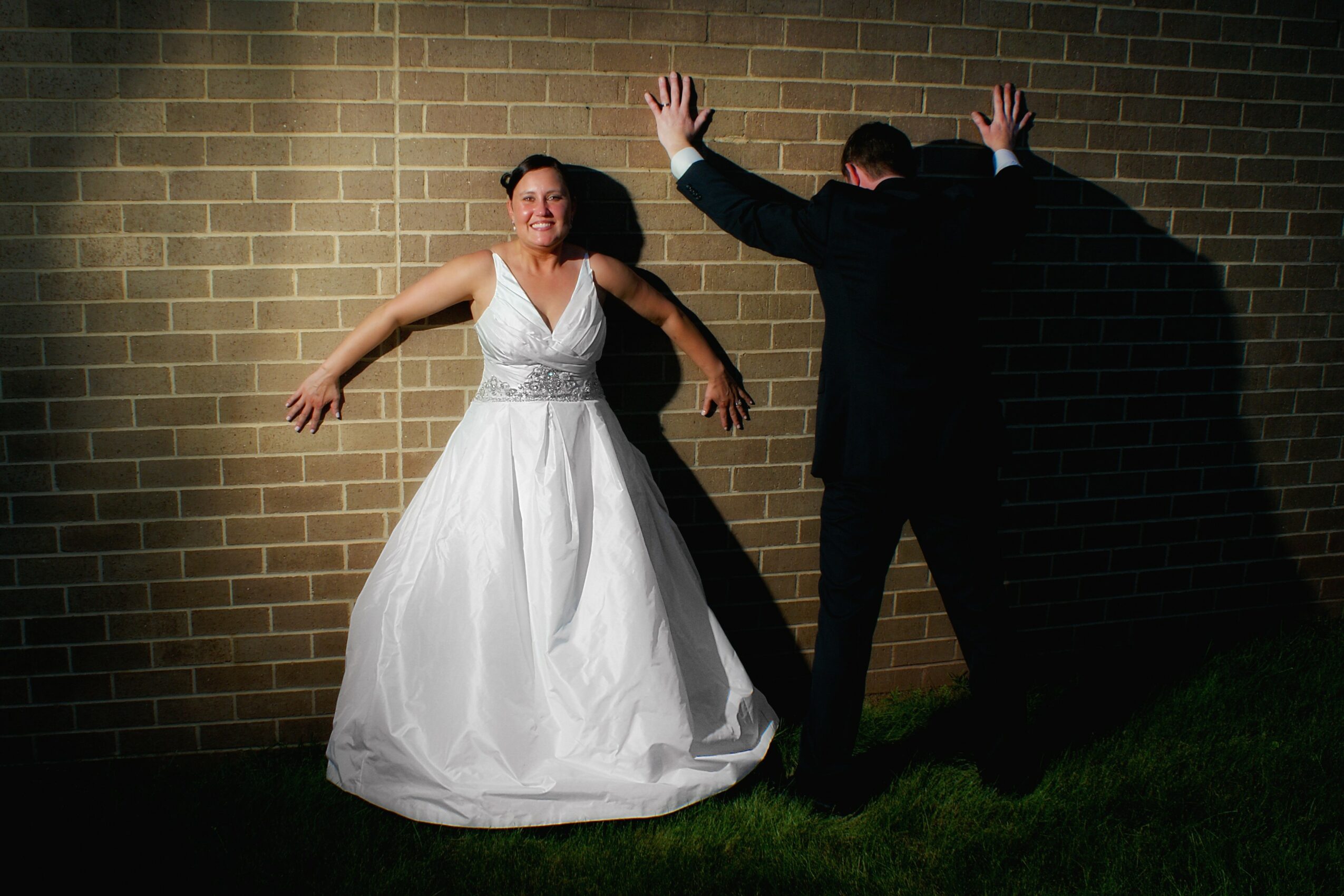 Woman in wedding dress smiling against brick wall, man in dark suit with hands on wall, shadow cast on wall