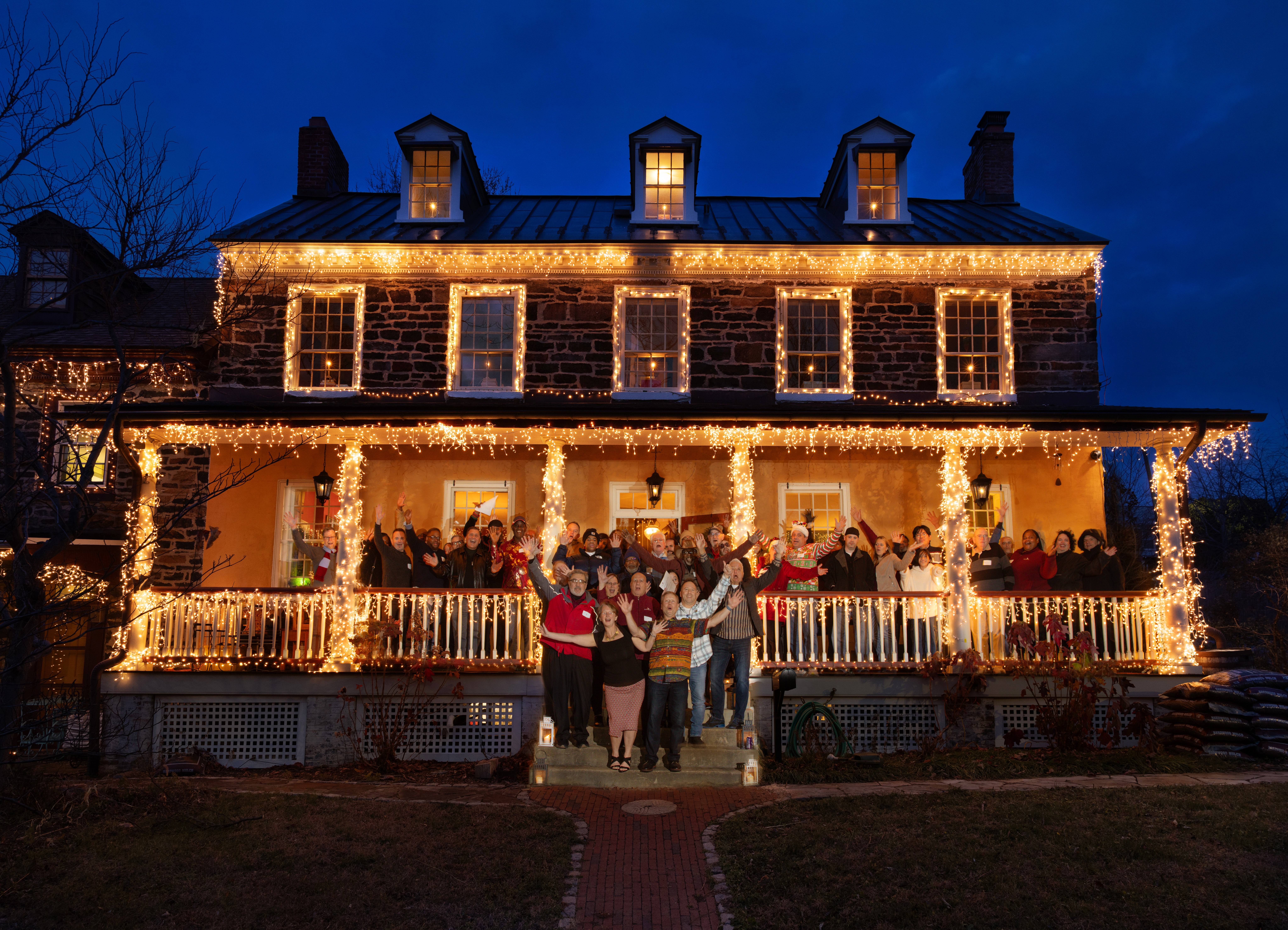 People gather on a decorated porch with Christmas lights in front of a two-story house at night.
