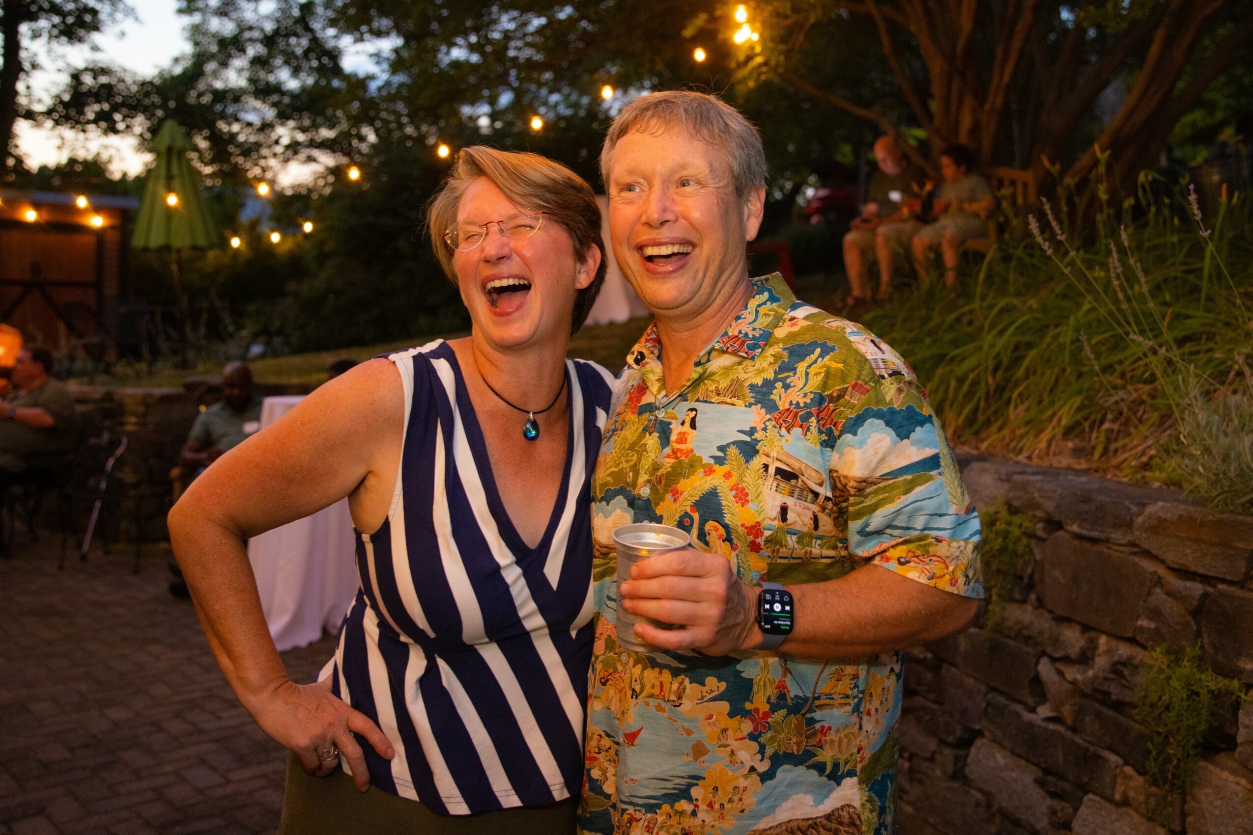 Two people laughing outdoors at night, one holding a drink, surrounded by trees and stone wall.