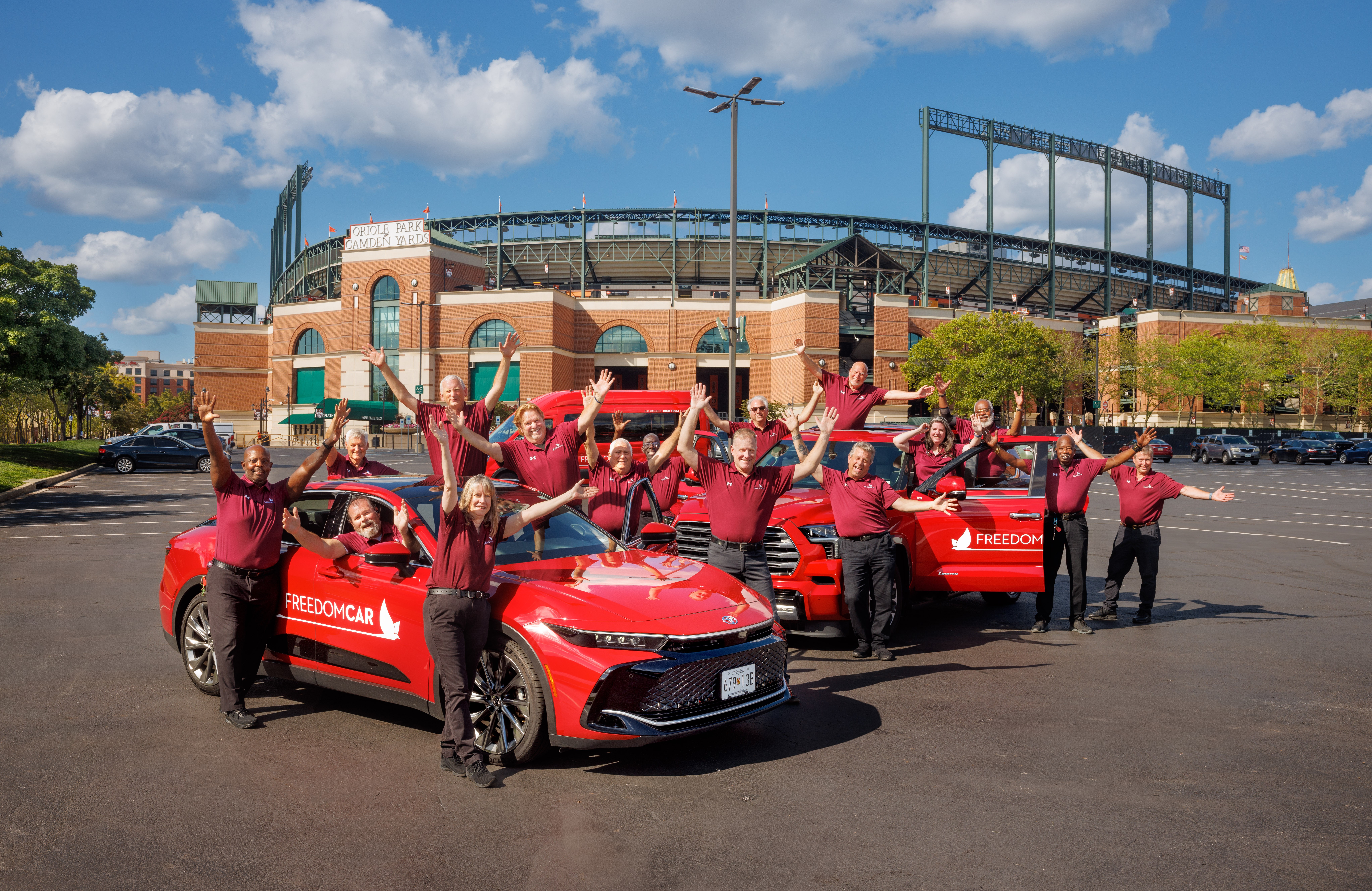 Group of people in red shirts posing with a red car in a parking lot outside a stadium, blue sky above.