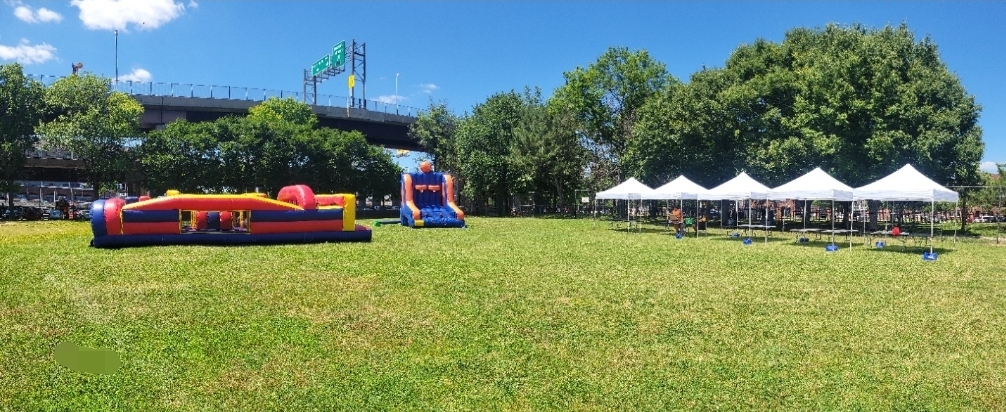 Open grassy field with inflatable bounce houses, tents, and trees under a blue sky.