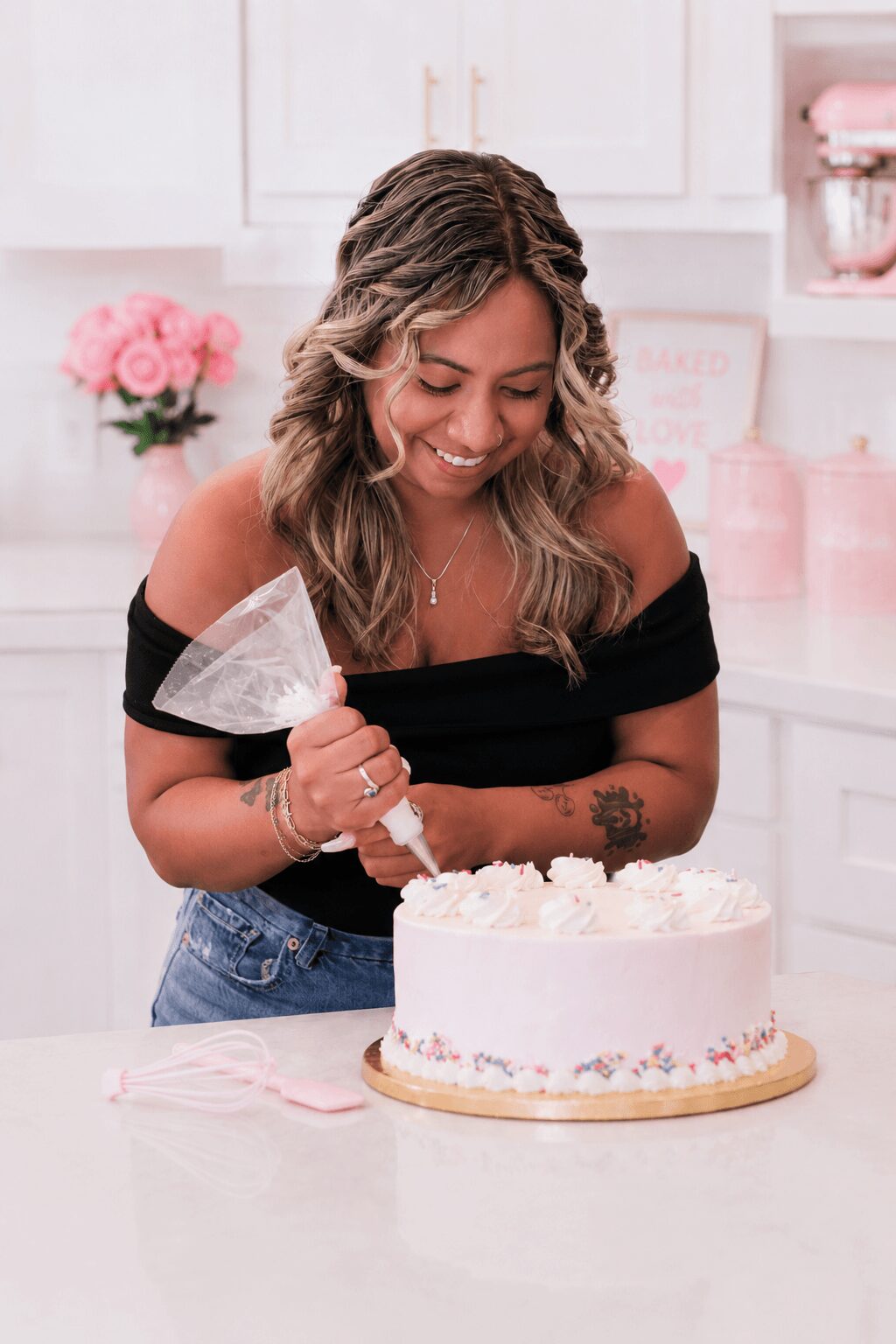 Woman with wavy hair smiles while decorating a cake with a spatula in a pink-themed kitchen.