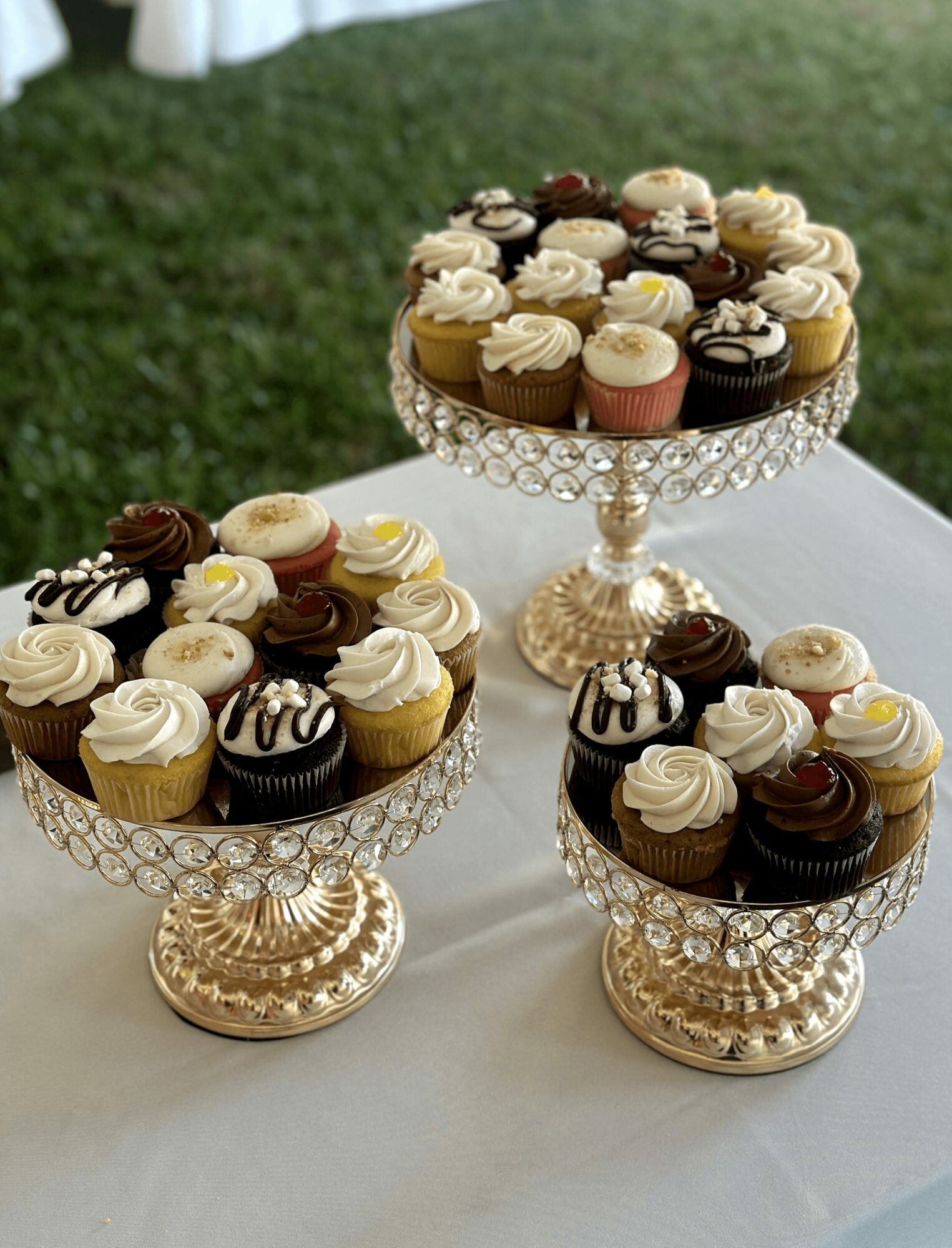 Three ornate glass cake stands with cupcakes on a white table outdoors, grass in the background.