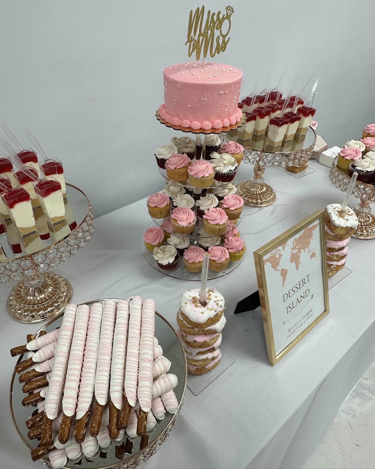 Dessert table with pink cake, cupcakes, and other sweets on glass stands, with a sign reading 'Dessert Table'.