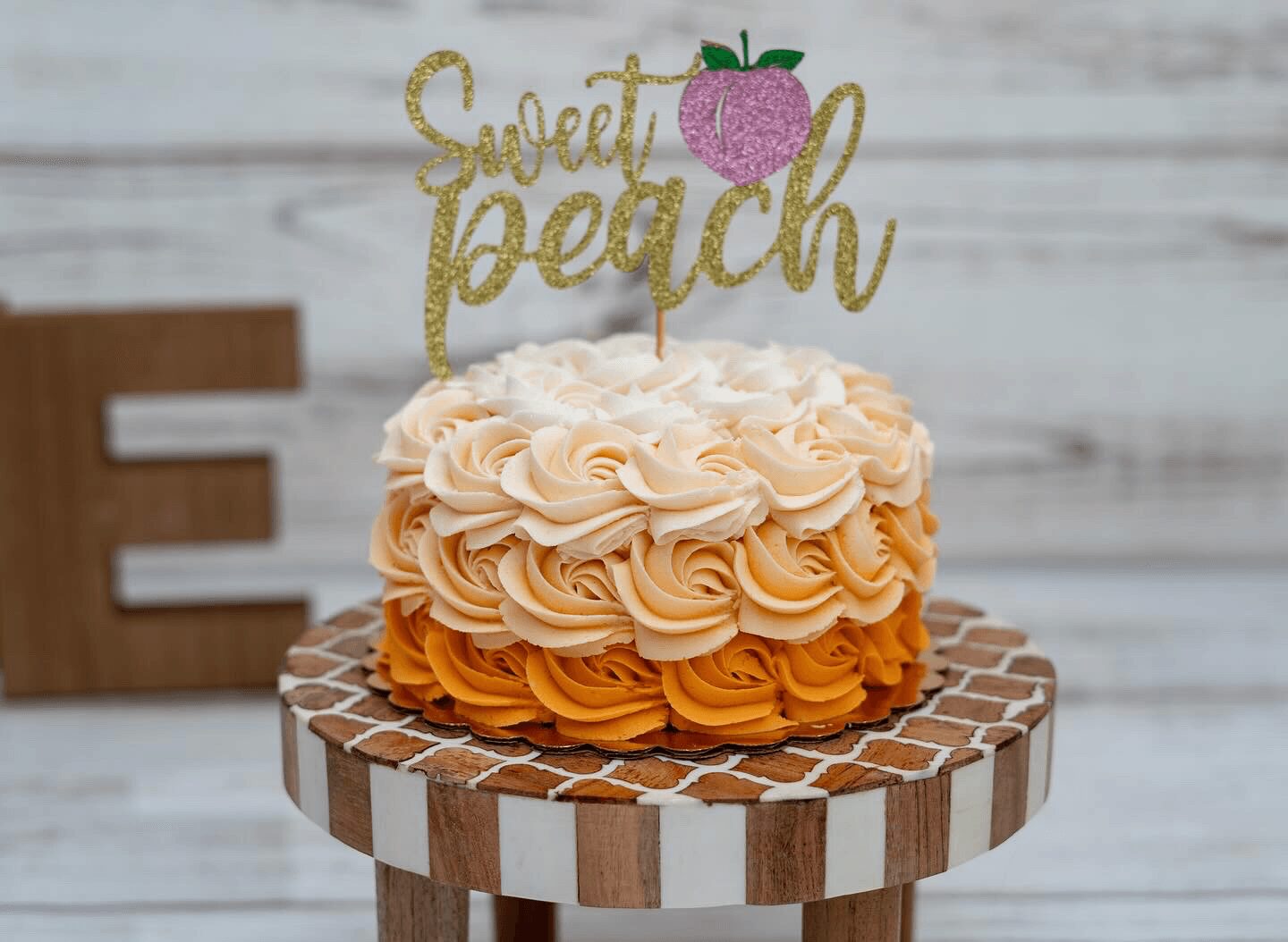 Decorative cake with peach-colored frosting on a wooden stand, topped with a glittery gold sign reading 'Sweet peach' and a pink fruit illustration.
