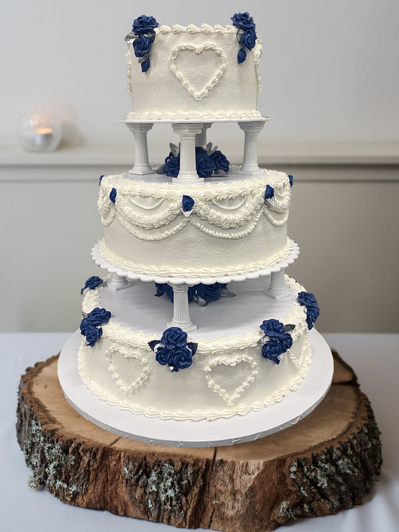 Three-tiered white wedding cake with blue flowers and heart-shaped decorations, on a wooden cake stand.