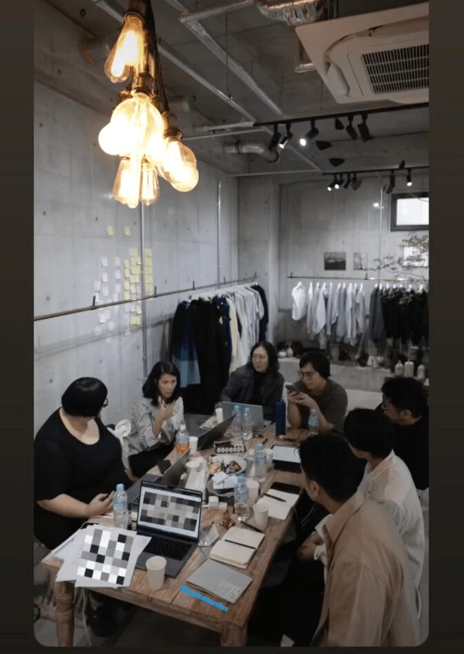 People sit around a table in a clothing store, with racks of clothes and a concrete wall in the background.