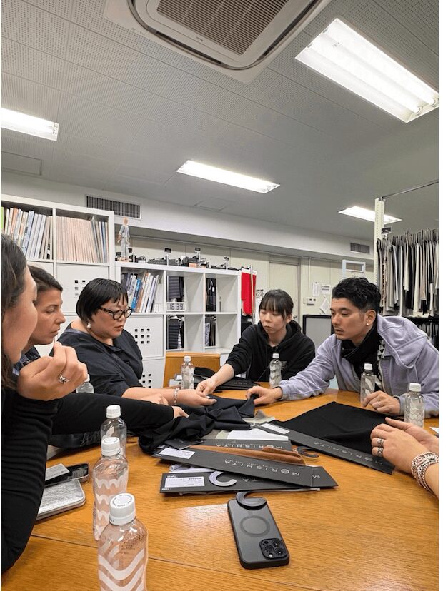 Group of people sitting around a table in a meeting room, engaging in discussion, with bookshelves and office supplies in background.