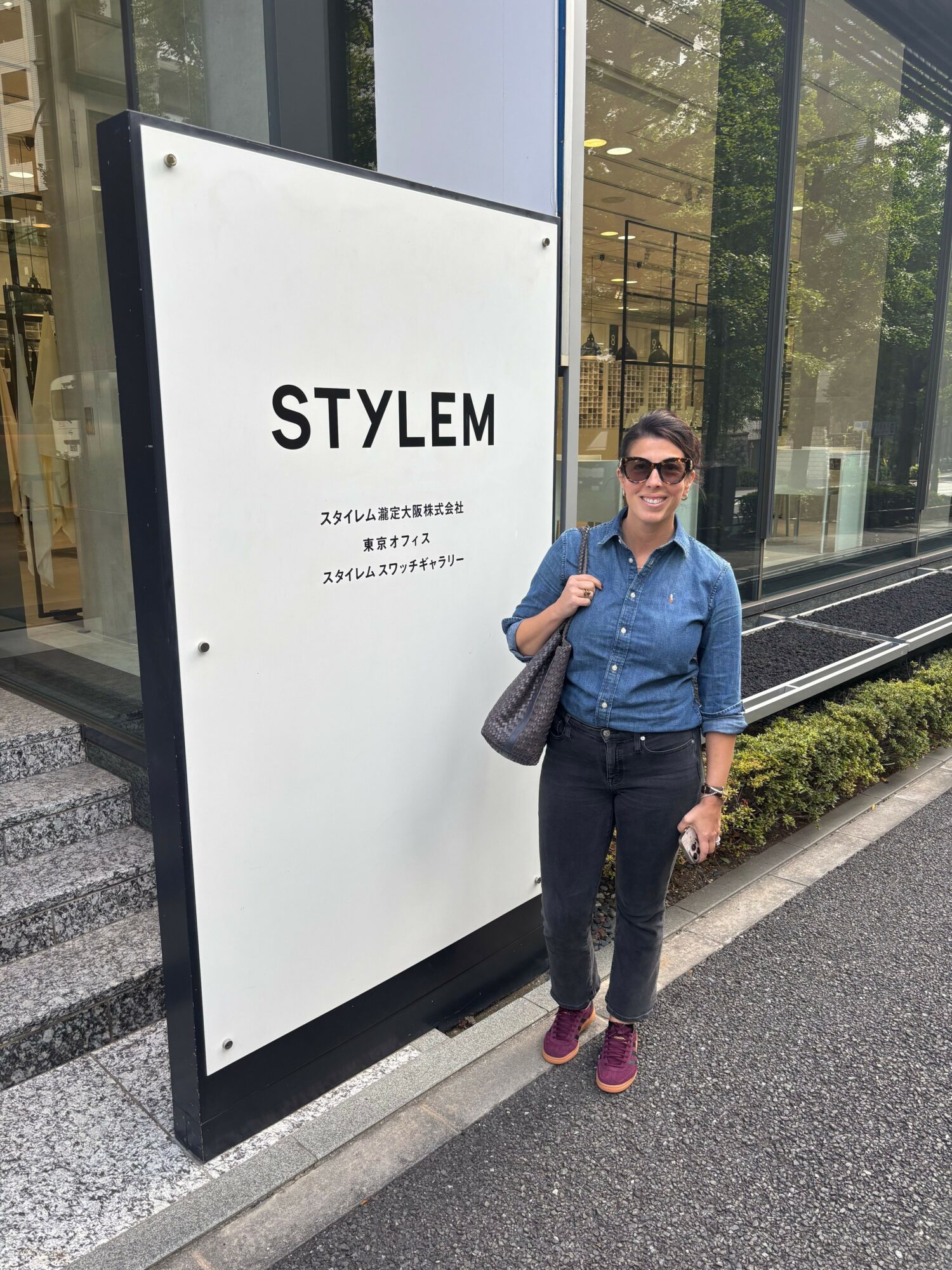 Woman standing next to a large white sign with black text, wearing sunglasses, a blue jacket, black pants, and purple shoes.